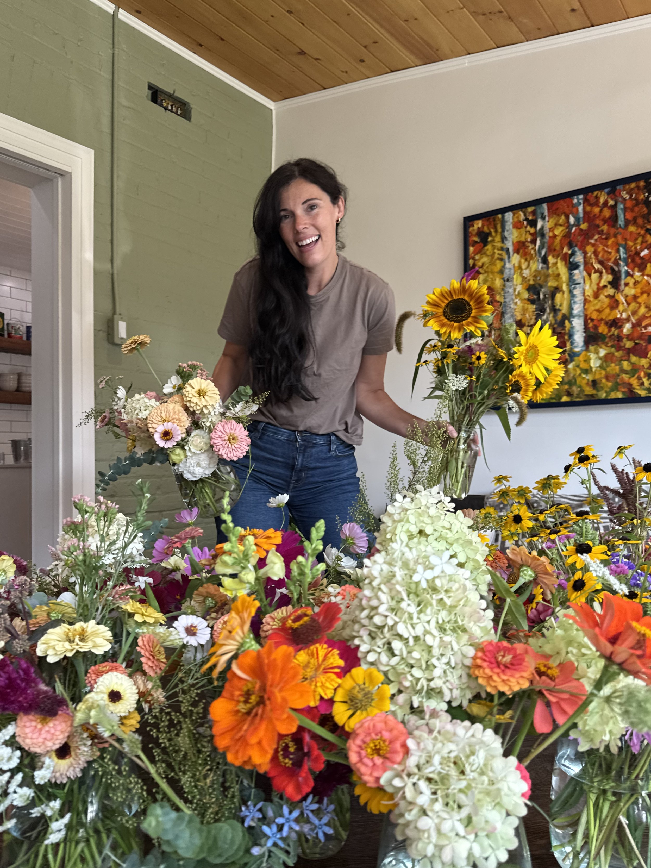 A woman with long dark hair smiling while arranging colorful flowers in a room with white and green walls, a wooden ceiling, and a large painting of a forest scene in the background.