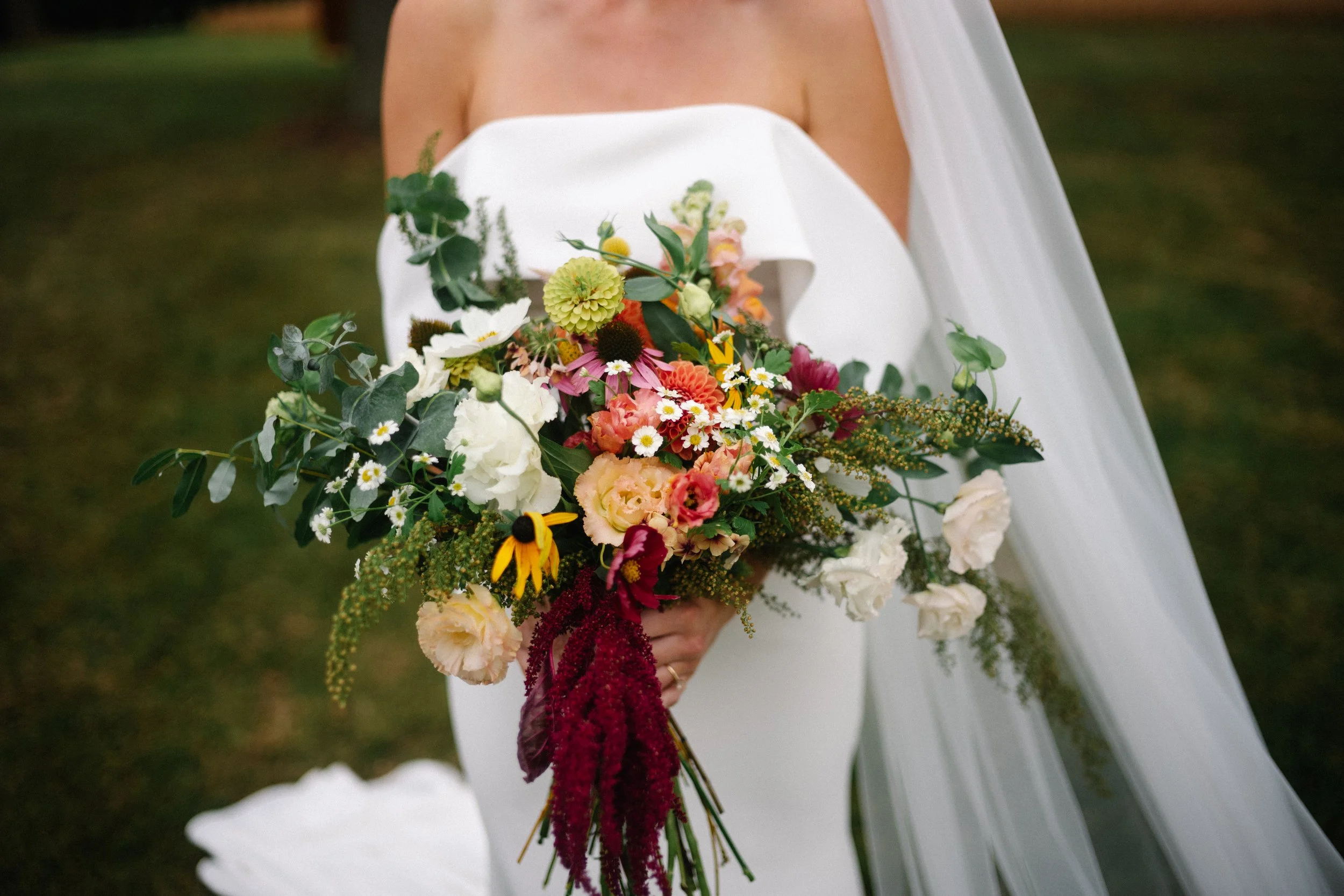 A bride holding a colorful bouquet of flowers, including daisies, sunflowers, and various greenery, with a white wedding dress and veil.