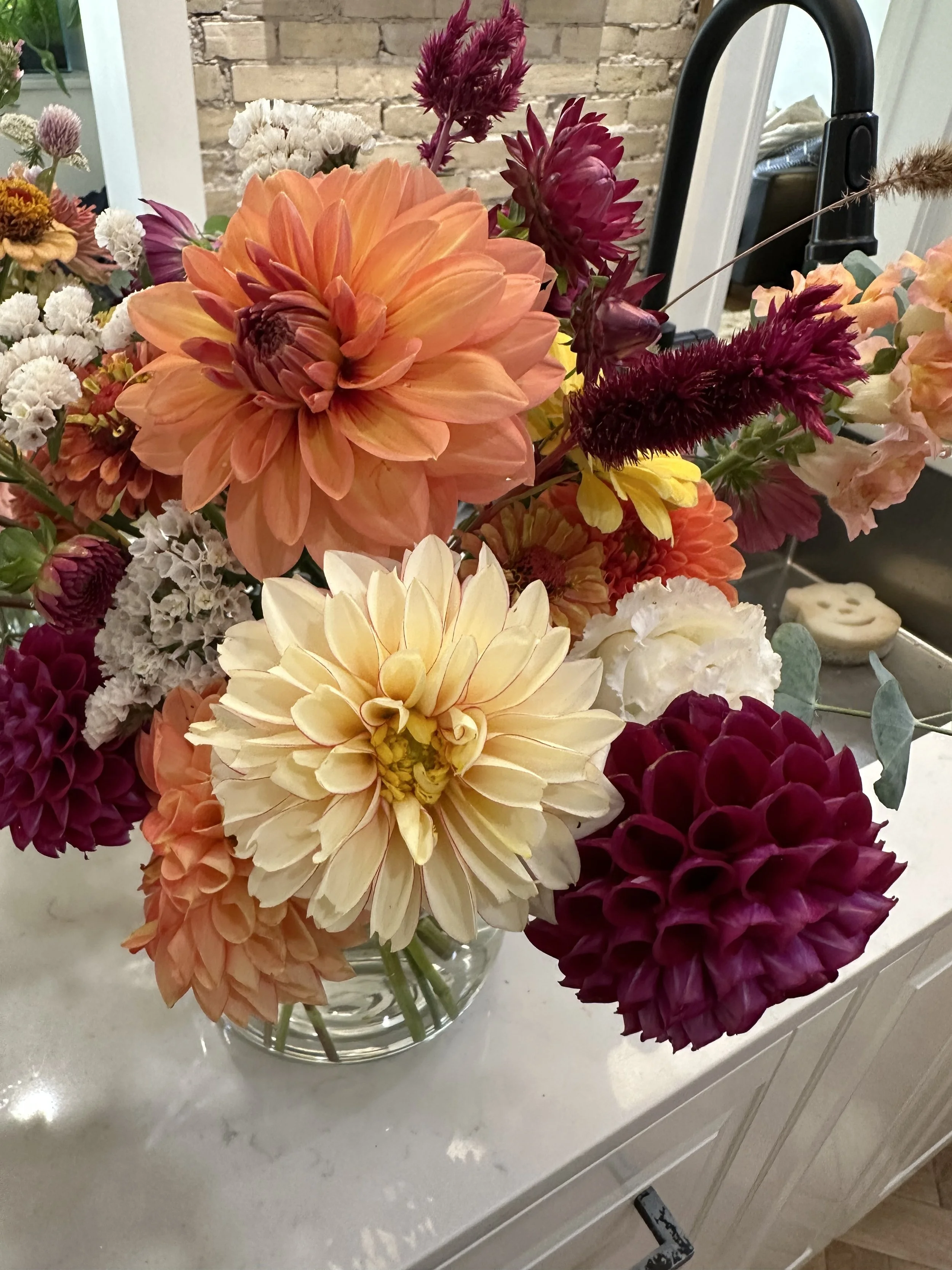 A bouquet of colorful flowers in a glass vase on a white countertop, with a brick wall and kitchen sink in the background.