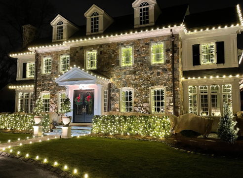 A large, elegant house decorated with Christmas lights at night, with a well-maintained lawn, potted plants, and pink flowers on the front porch.