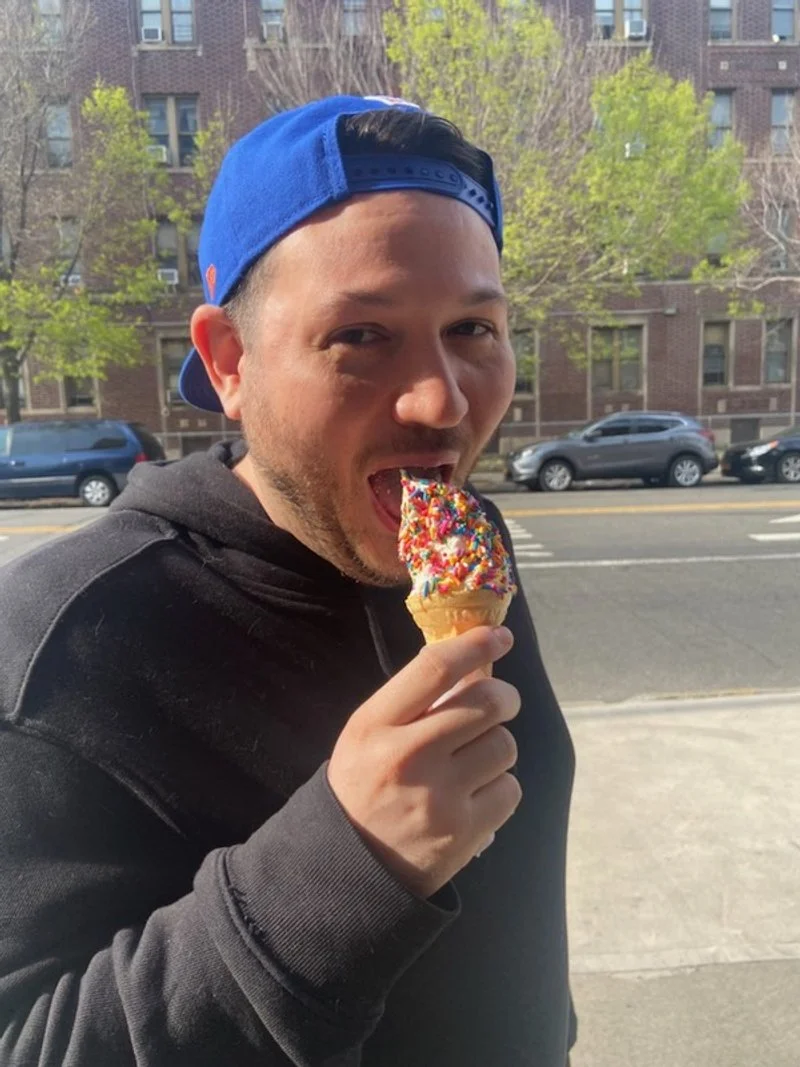 A man wearing a blue cap and black hoodie, standing outdoors on a city street, is about to eat a colorful ice cream cone with sprinkles.
