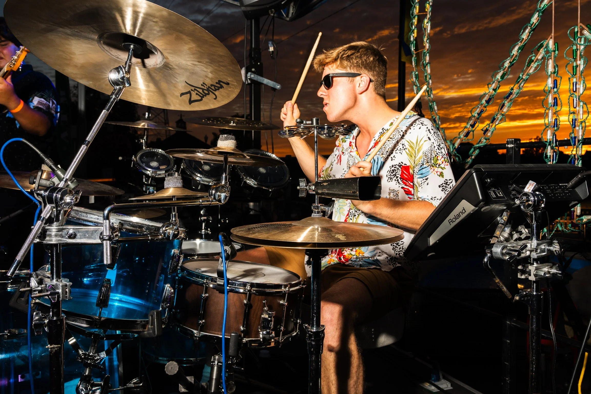 A young man wearing sunglasses and a colorful floral shirt playing a drum set outdoors during sunset, with chains and sunset sky in the background.