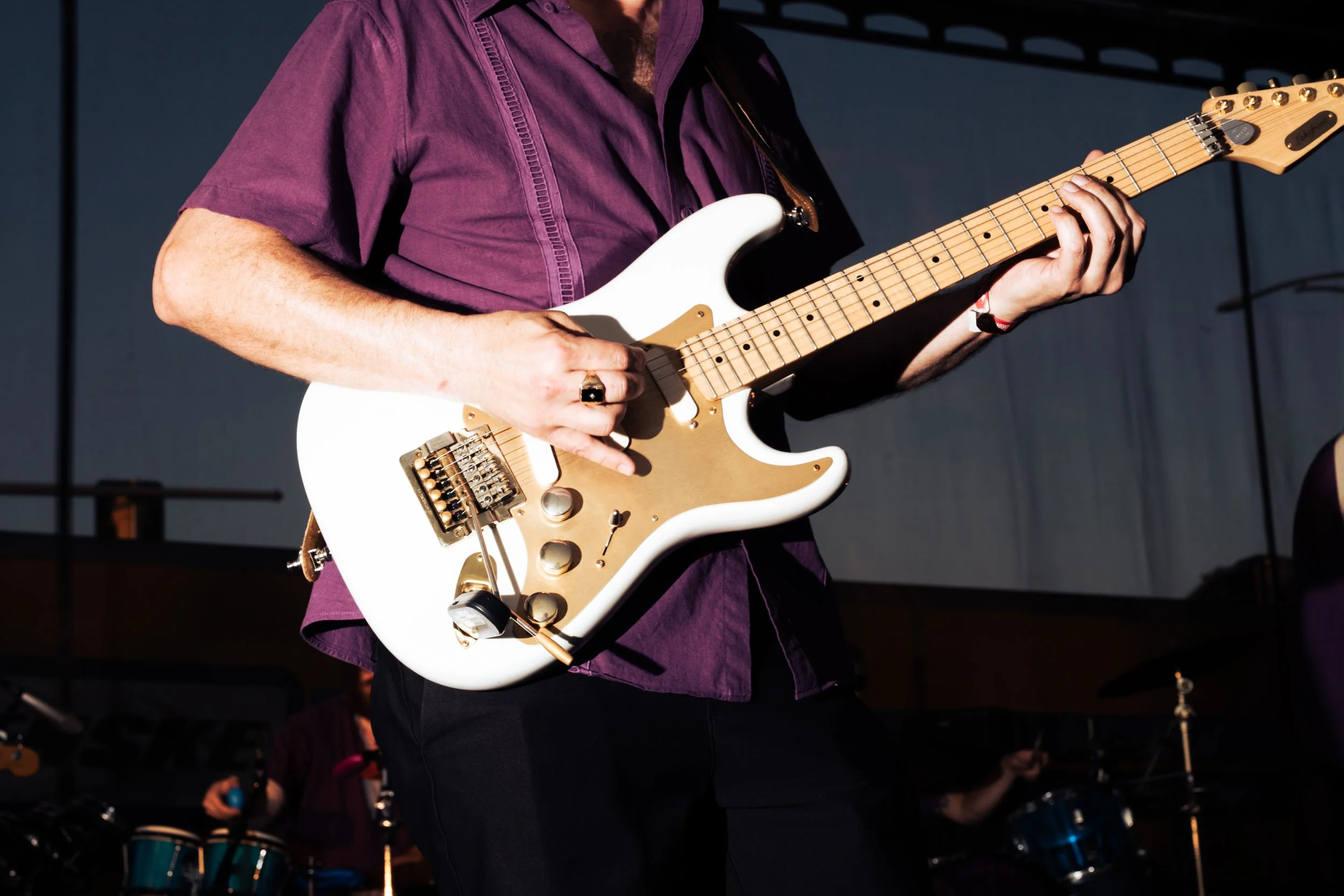 Close-up of a person playing a white electric guitar with gold hardware, wearing a purple shirt, on stage with a drum set in the background.