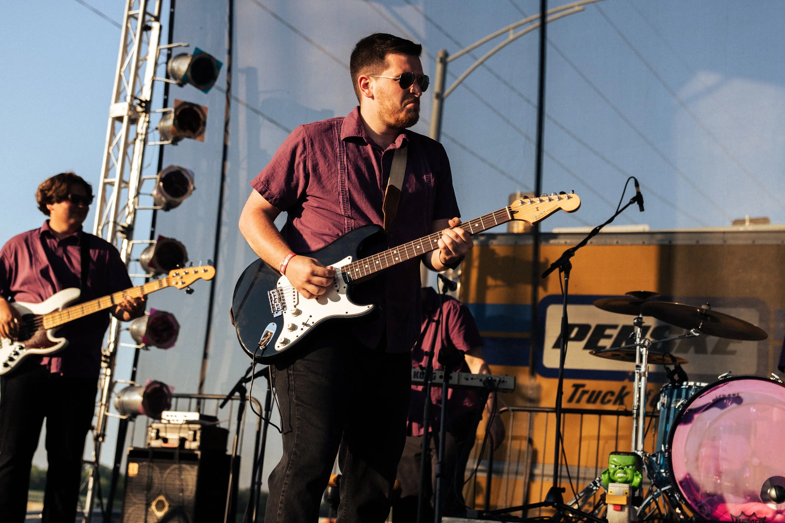 Musicians performing live on outdoor stage, playing electric guitars and keyboard, with stage lighting and equipment nearby.