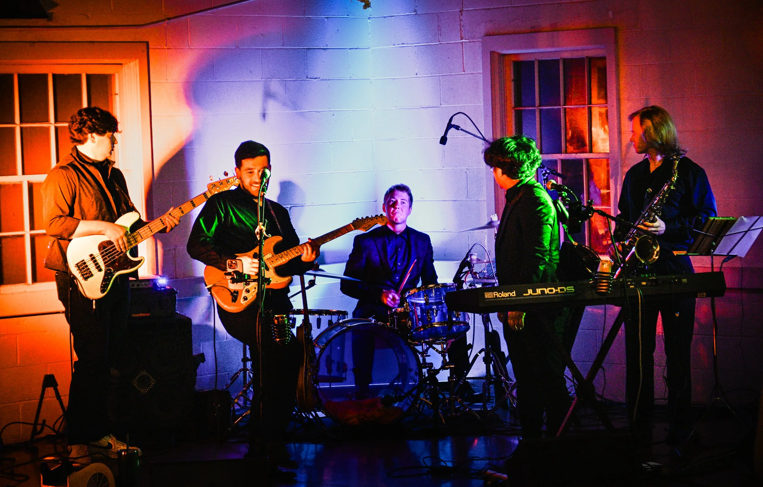 A five-member band performing on stage with each member playing a different instrument, illuminated by colorful stage lights.