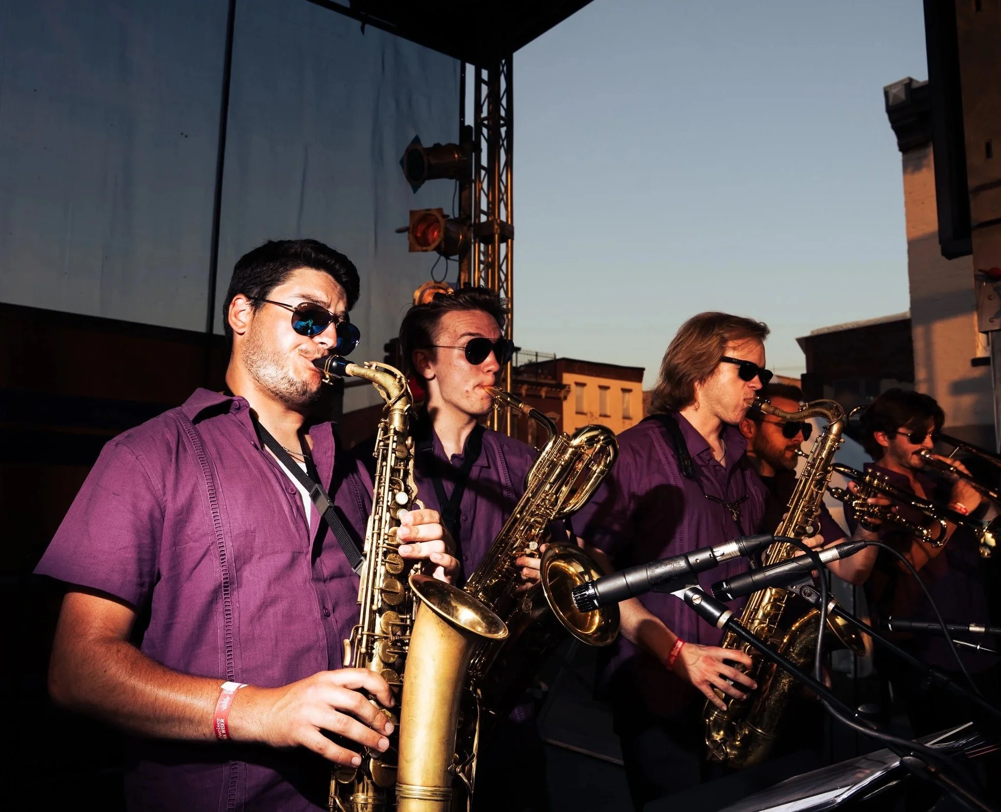 Six musicians playing saxophones on an outdoor stage during sunset, wearing matching purple shirts and sunglasses.