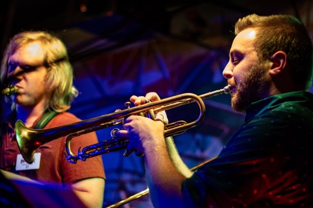 Two musicians playing brass instruments, one with blonde hair and glasses, the other with a beard, in a dimly lit setting with colorful lighting.