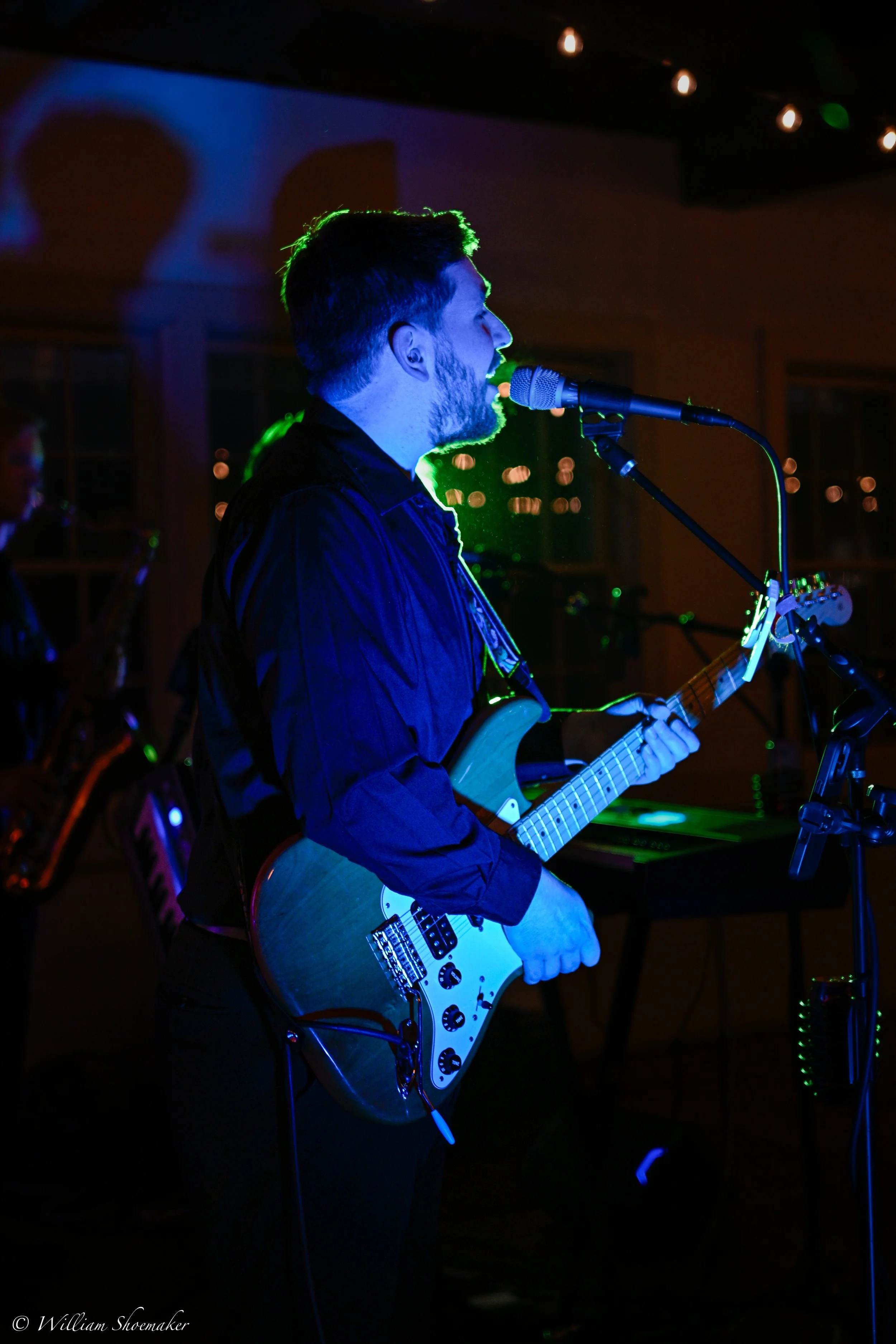 A male musician in a dark shirt playing an electric guitar and singing into a microphone during a live performance, with colorful stage lighting and another band member playing keyboard in the background.