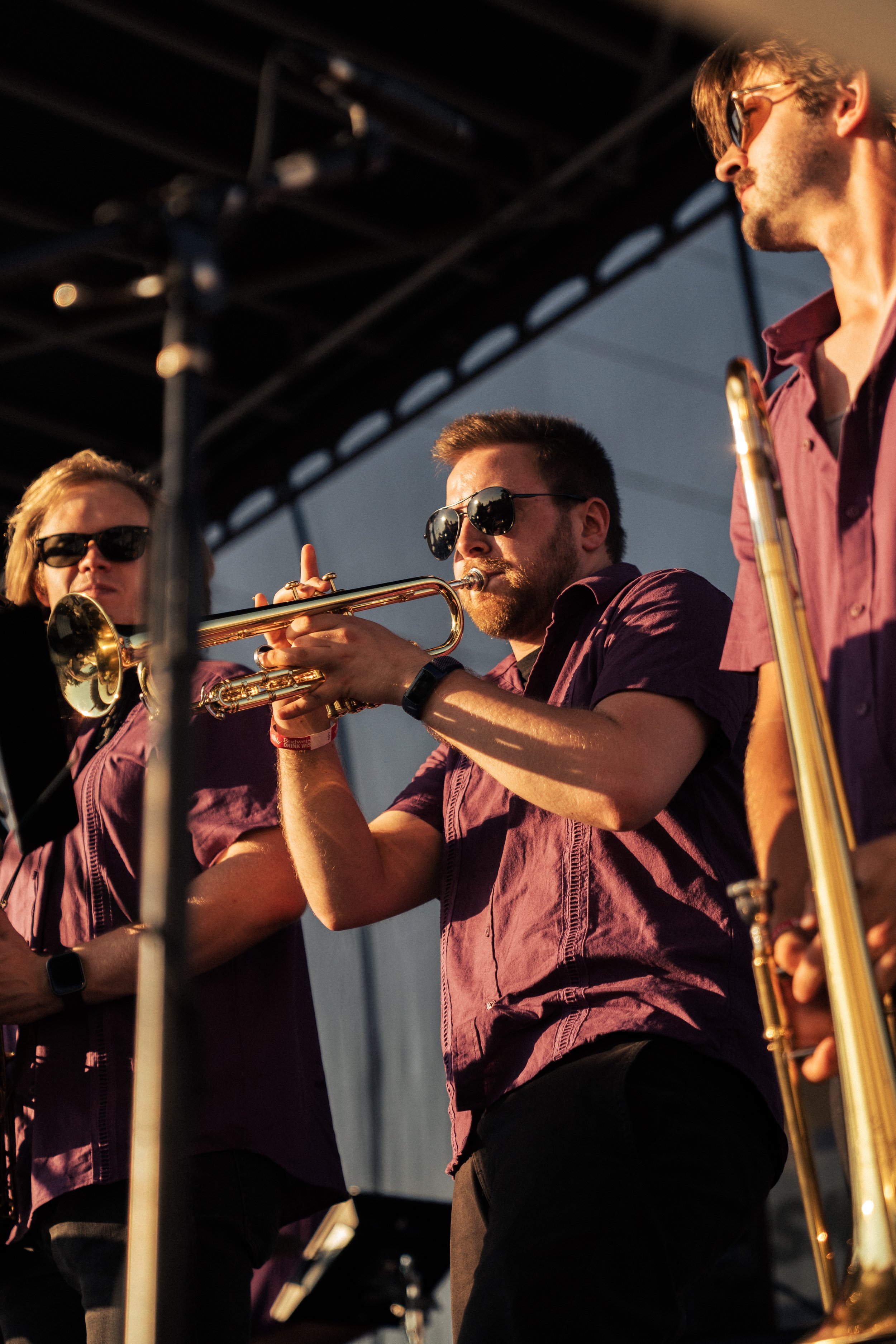 Three musicians playing brass instruments on stage during a performance, wearing purple shirts and sunglasses.