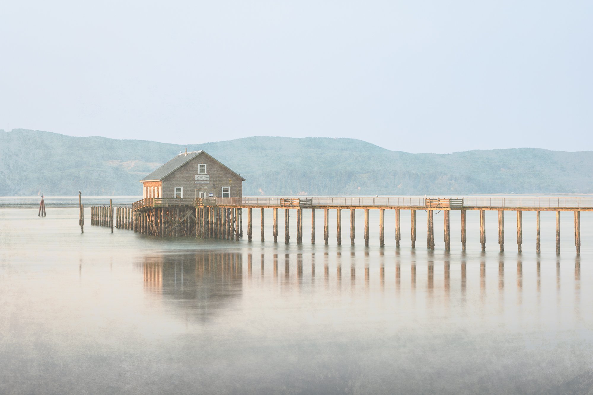 A wooden pier extending into calm water with a small wooden building on stilts at the end, mountains in the background, and a light, hazy sky.