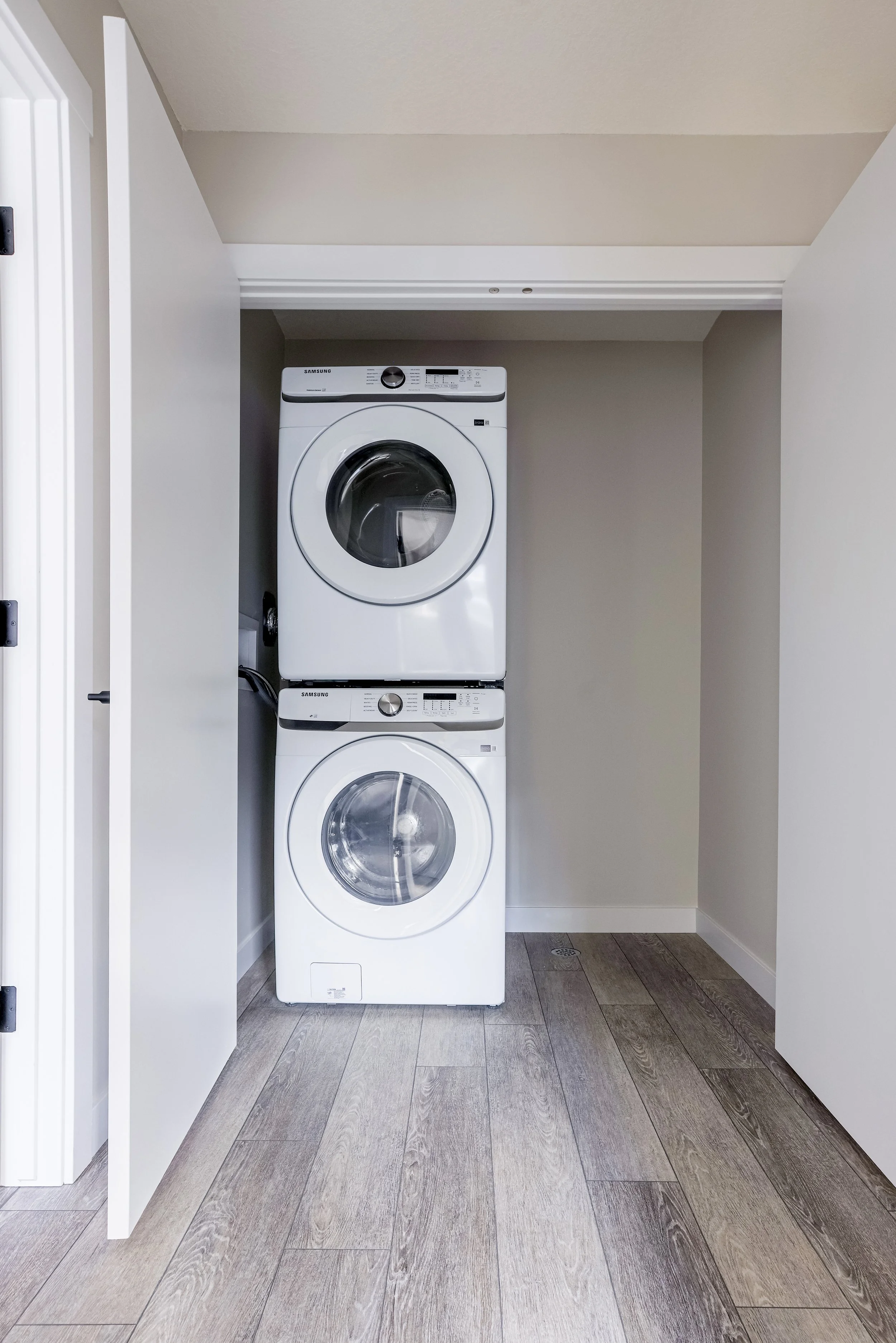 White stacked laundry washer and dryer inside a small closet with open white doors, wood flooring, and beige walls.