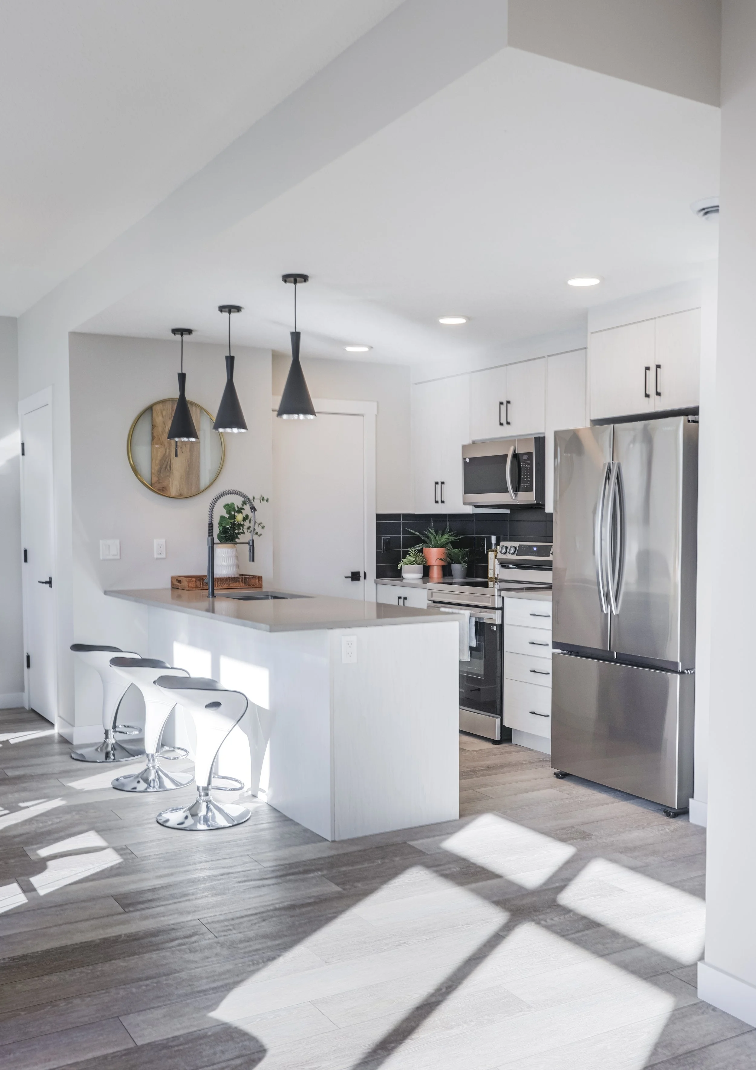 Modern kitchen with white cabinets, stainless steel appliances, black backsplash, and hardwood flooring. Features a kitchen island with three white barstools, black pendant lights, and natural sunlight streaming in through windows.