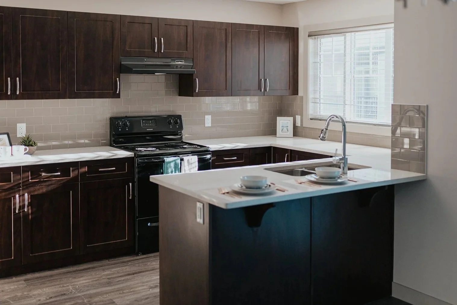 Modern kitchen with dark wood cabinets, white countertop, black stove, stainless steel sink, window with blinds, and decorative items.