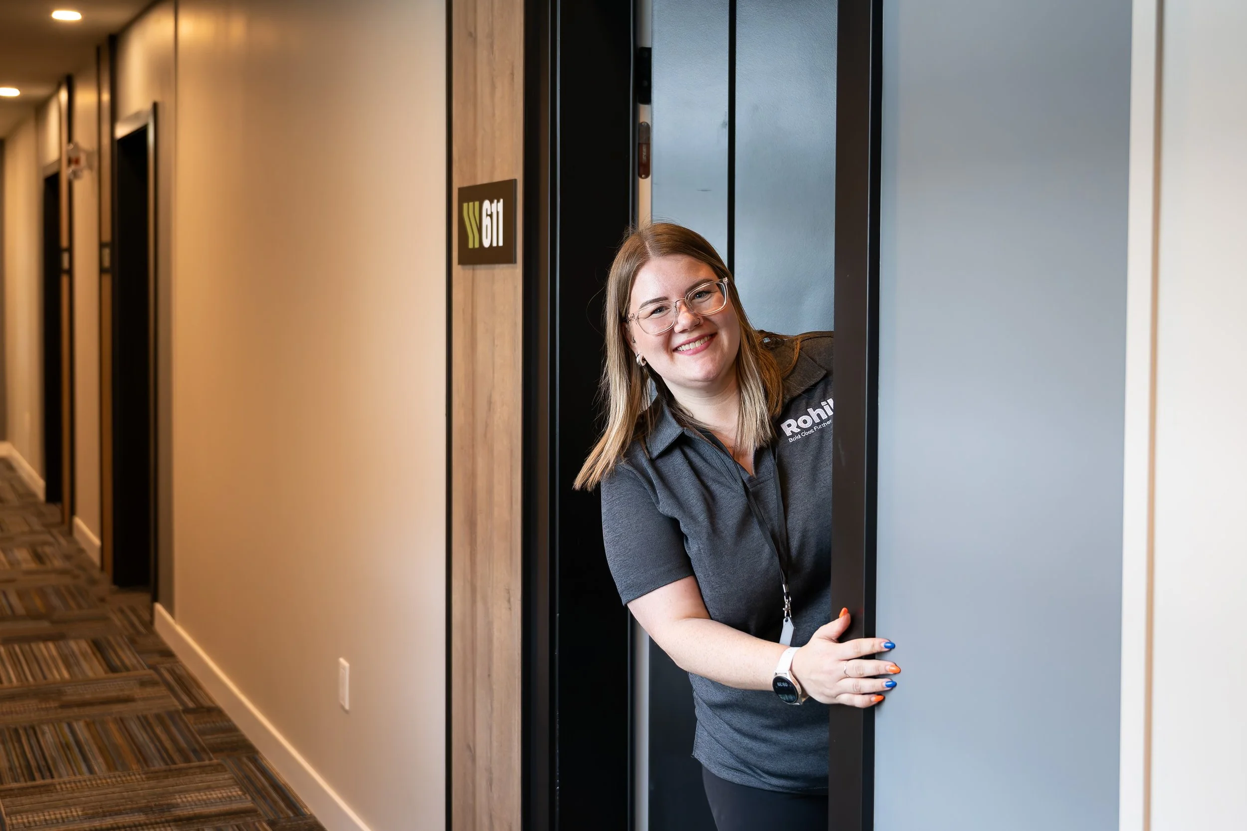 A smiling woman with glasses leaning out of an elevator door in a corridor of an office building.