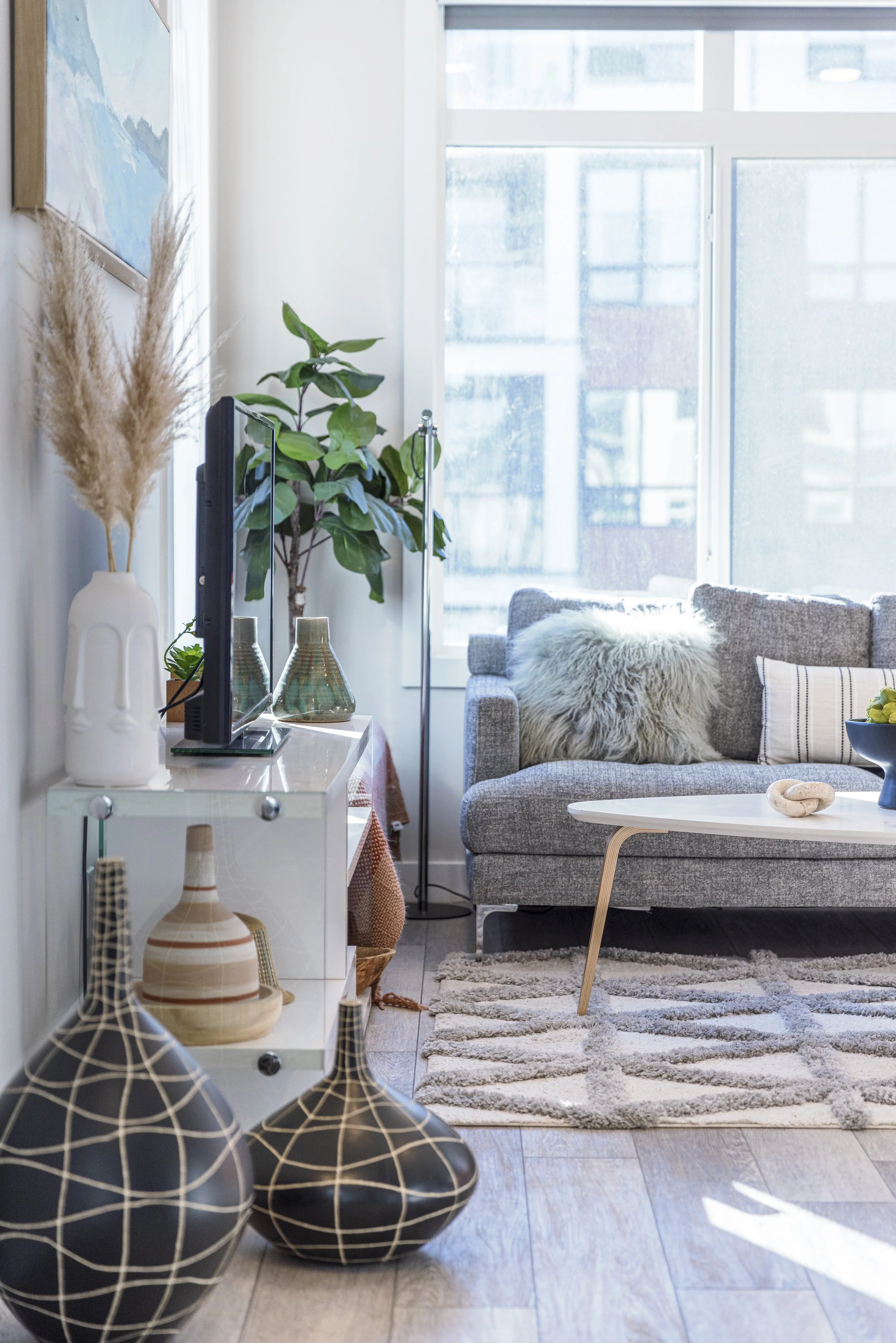 Living room with gray sofa, white coffee table, potted plant, TV on white stand, decorative vases, patterned rug, and large window with city view.