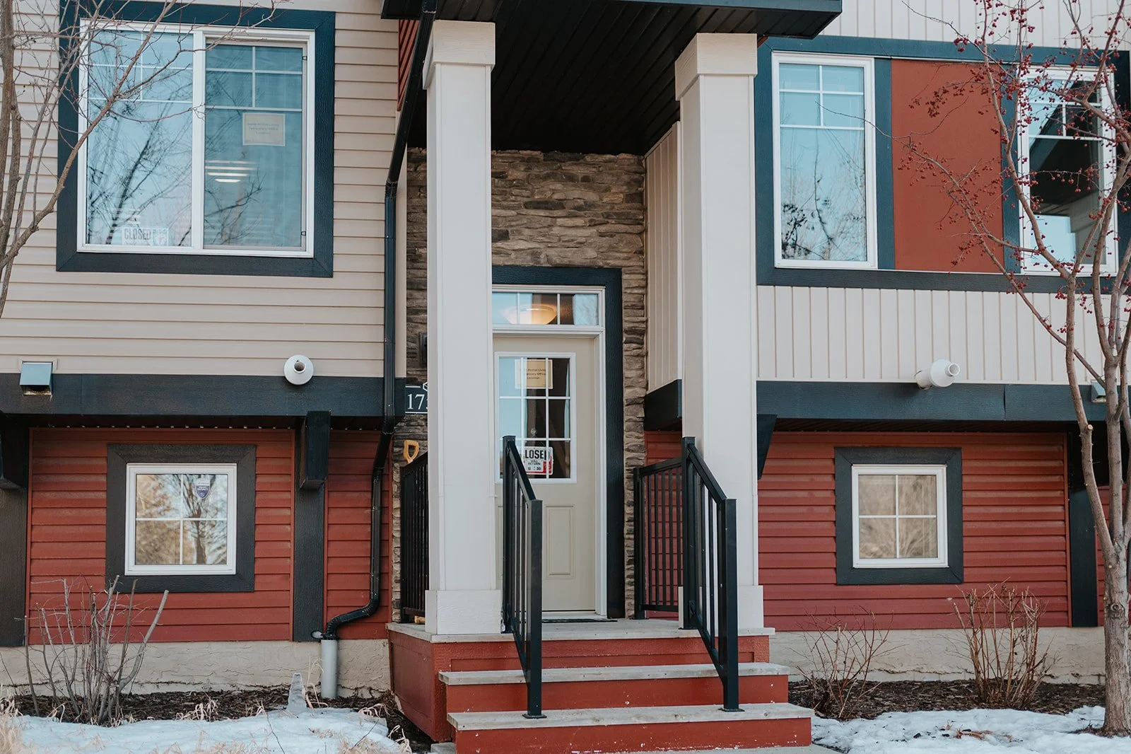 Modern two-story townhouse with beige, red, and blue siding, stone accents, and a small porch with steps and black railing. Snow on the ground and 