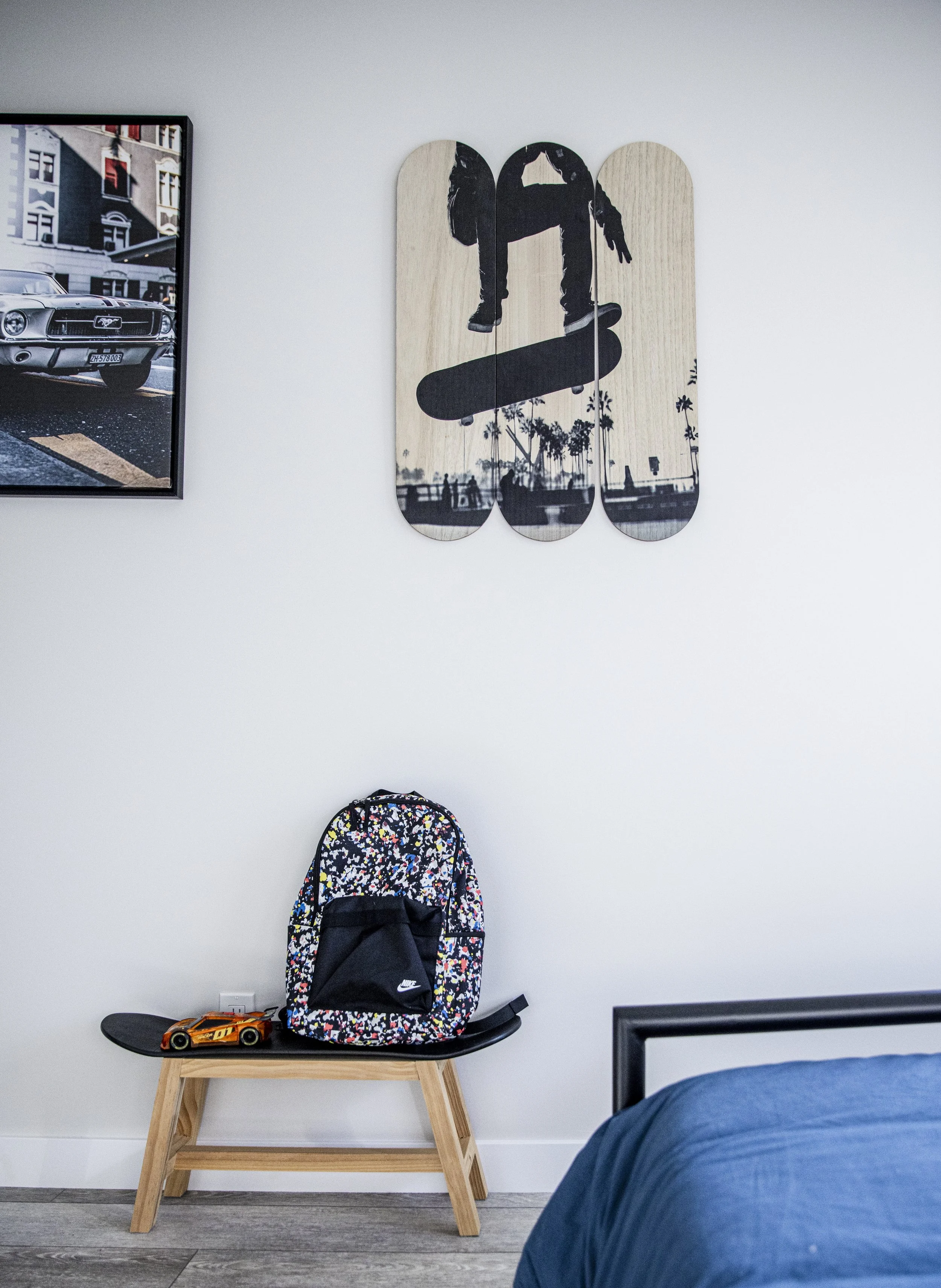 A bedroom corner with a skateboard, a colorful backpack, and a toy car on a wooden stool. A black and white skateboard-themed wall art hangs above, with part of a framed photo of a classic car and city street scene visible nearby.