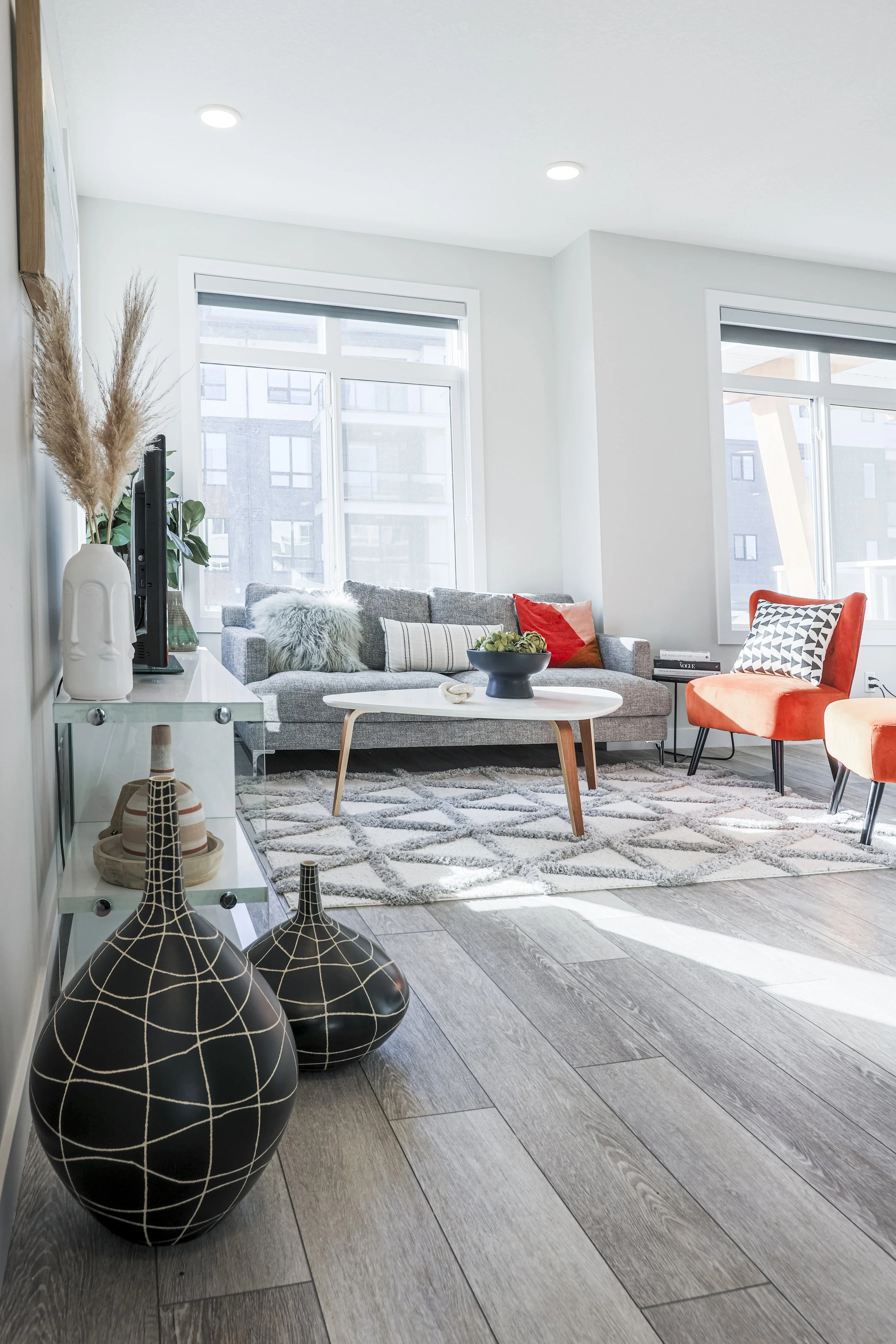 Modern living room with large windows, gray couch with decorative pillows, orange accent chairs, a white coffee table, and decorative vases with pampas grass and pottery.
