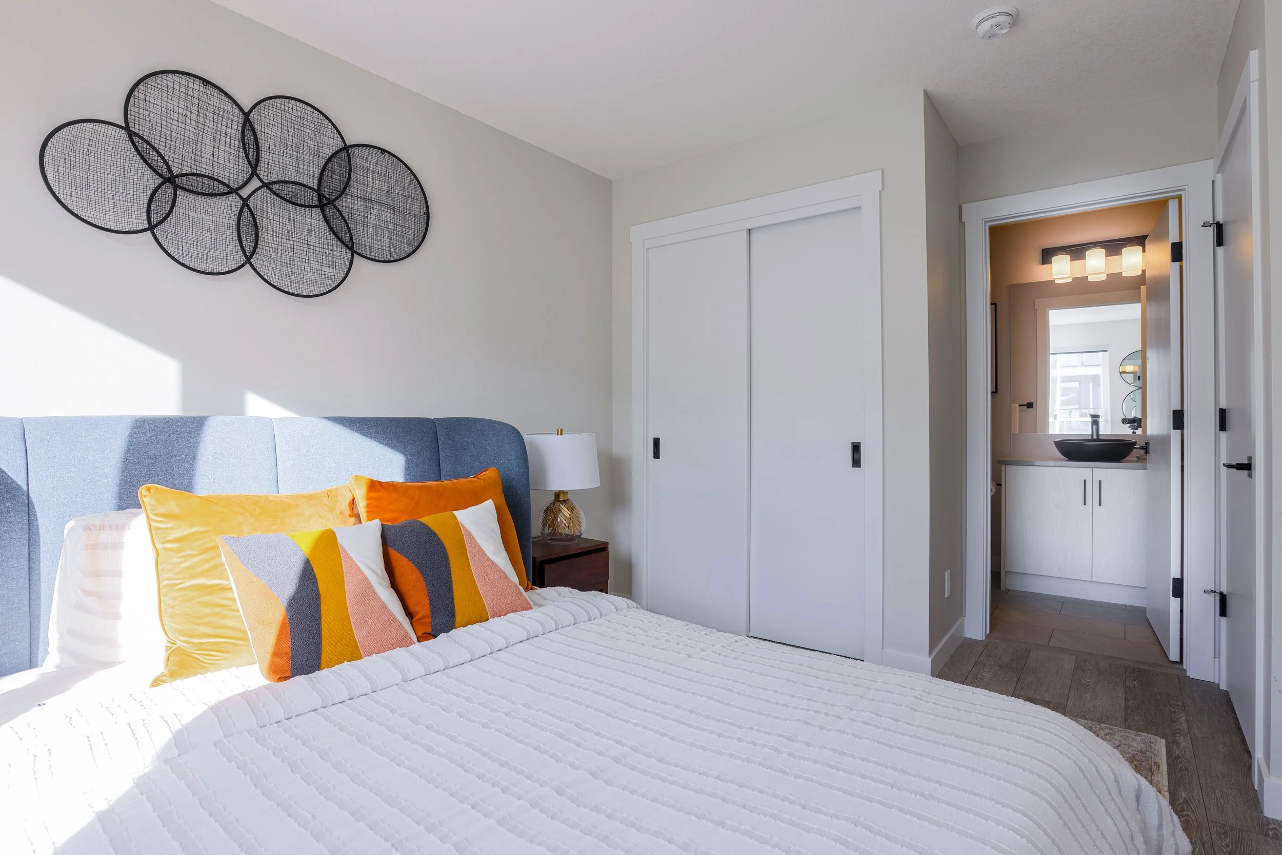 A bedroom with a bed that has a blue headboard and colorful pillows. There is a nightstand with a lamp. The room has white walls and a closet with sliding doors. An open door reveals a bathroom with a vanity, round black sink, and multiple lights abo