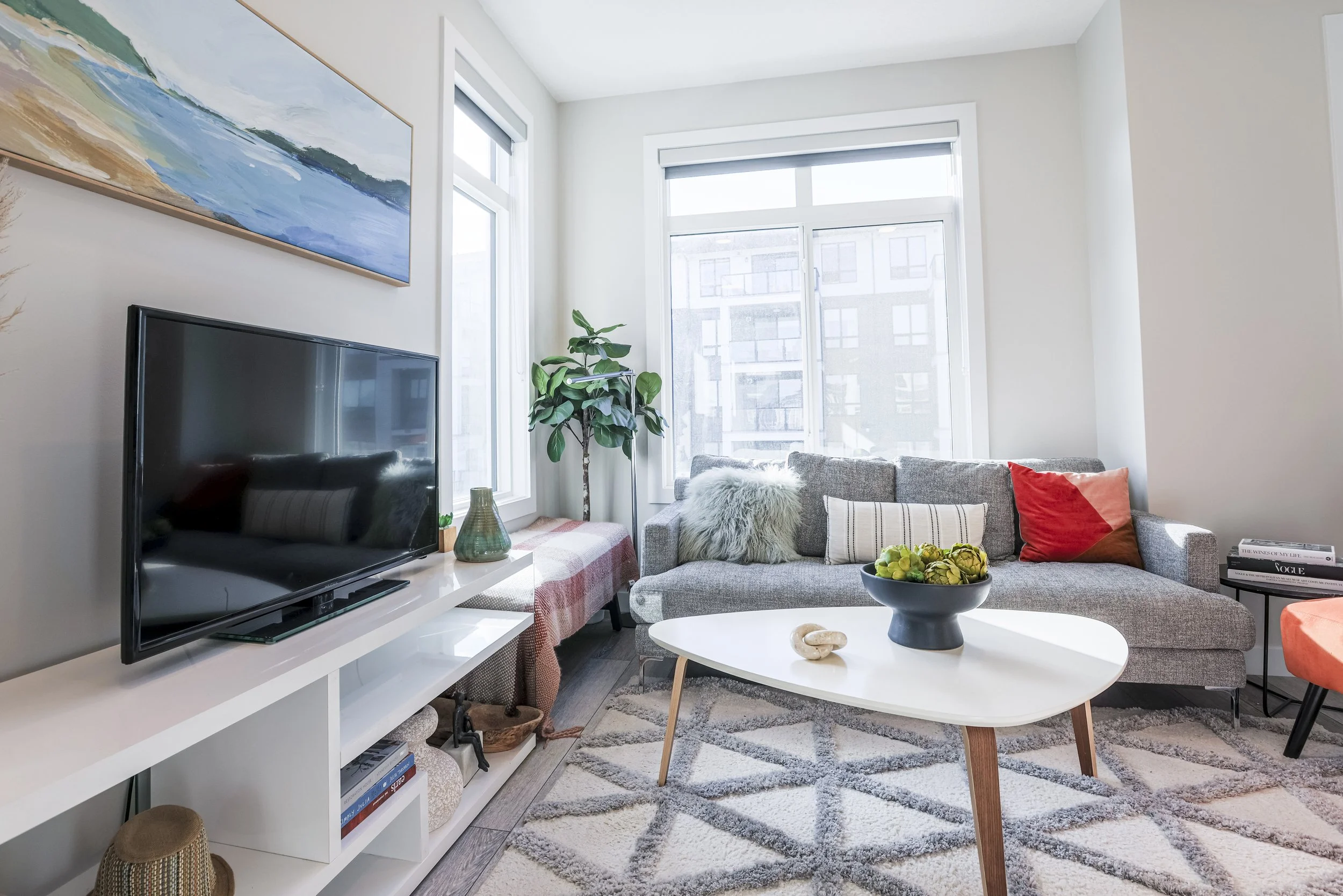 Bright living room with a gray sofa, colorful pillows, a coffee table with a bowl of artichokes, a TV on a white shelf, a tall houseplant, and windows letting in natural light.