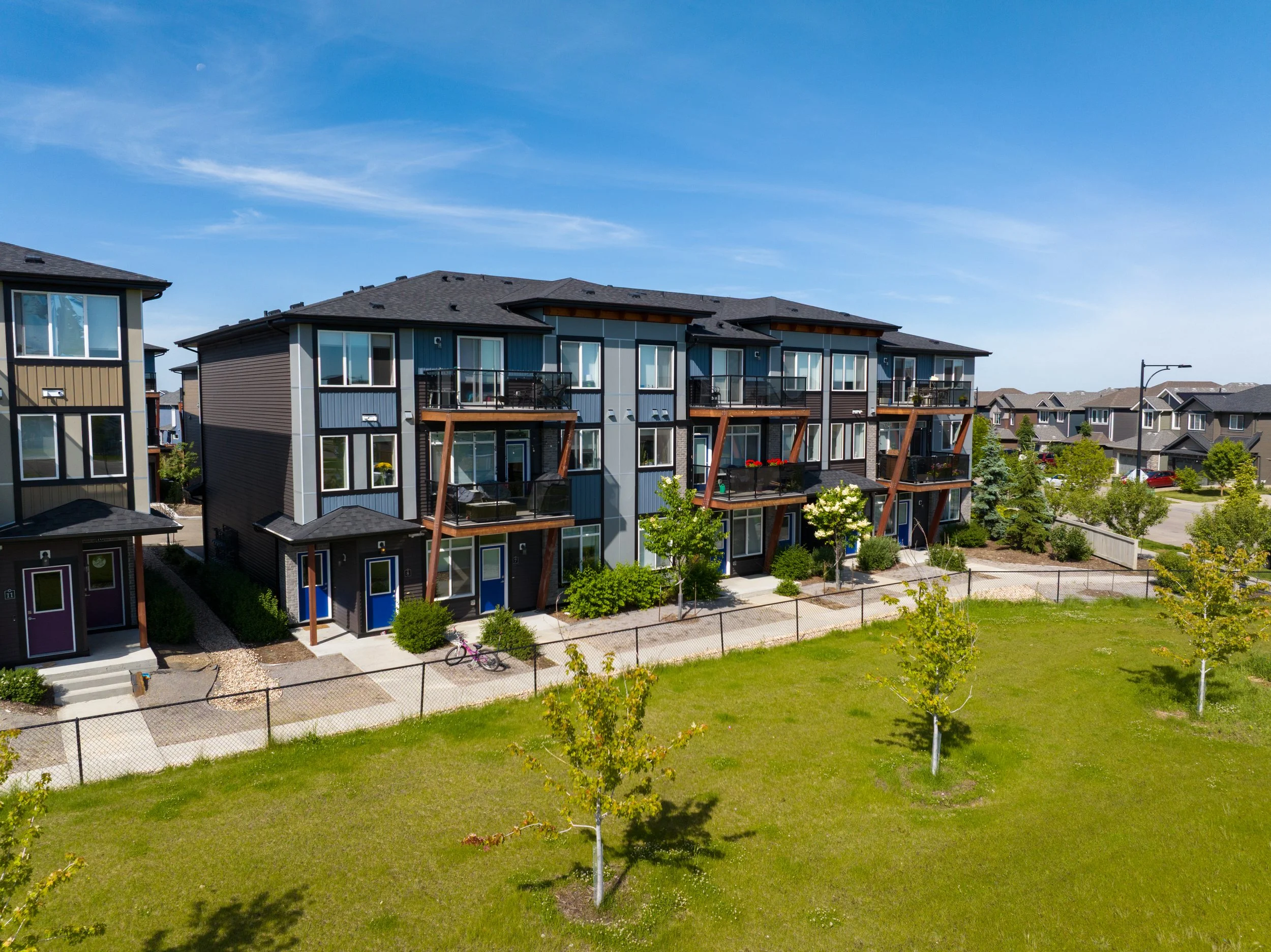 A residential neighborhood with modern multi-story apartment buildings featuring large windows and balconies, surrounded by green grass, young trees, and a clear blue sky with wispy clouds and a visible moon.
