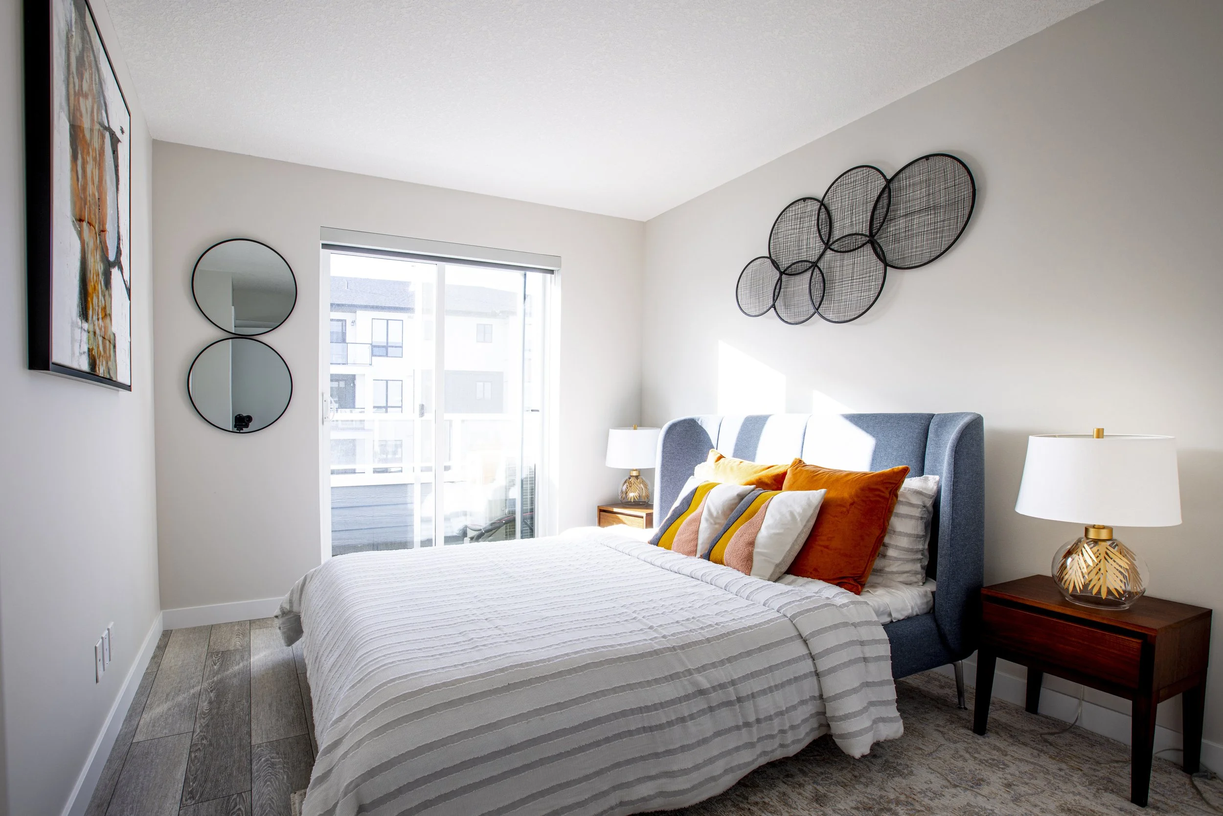 A bright bedroom with a bed dressed in white striped bedding and accented by orange and white pillows. Two matching white lamps with gold bases sit on dark wood nightstands on either side of the bed. The walls are decorated with abstract wall art, in