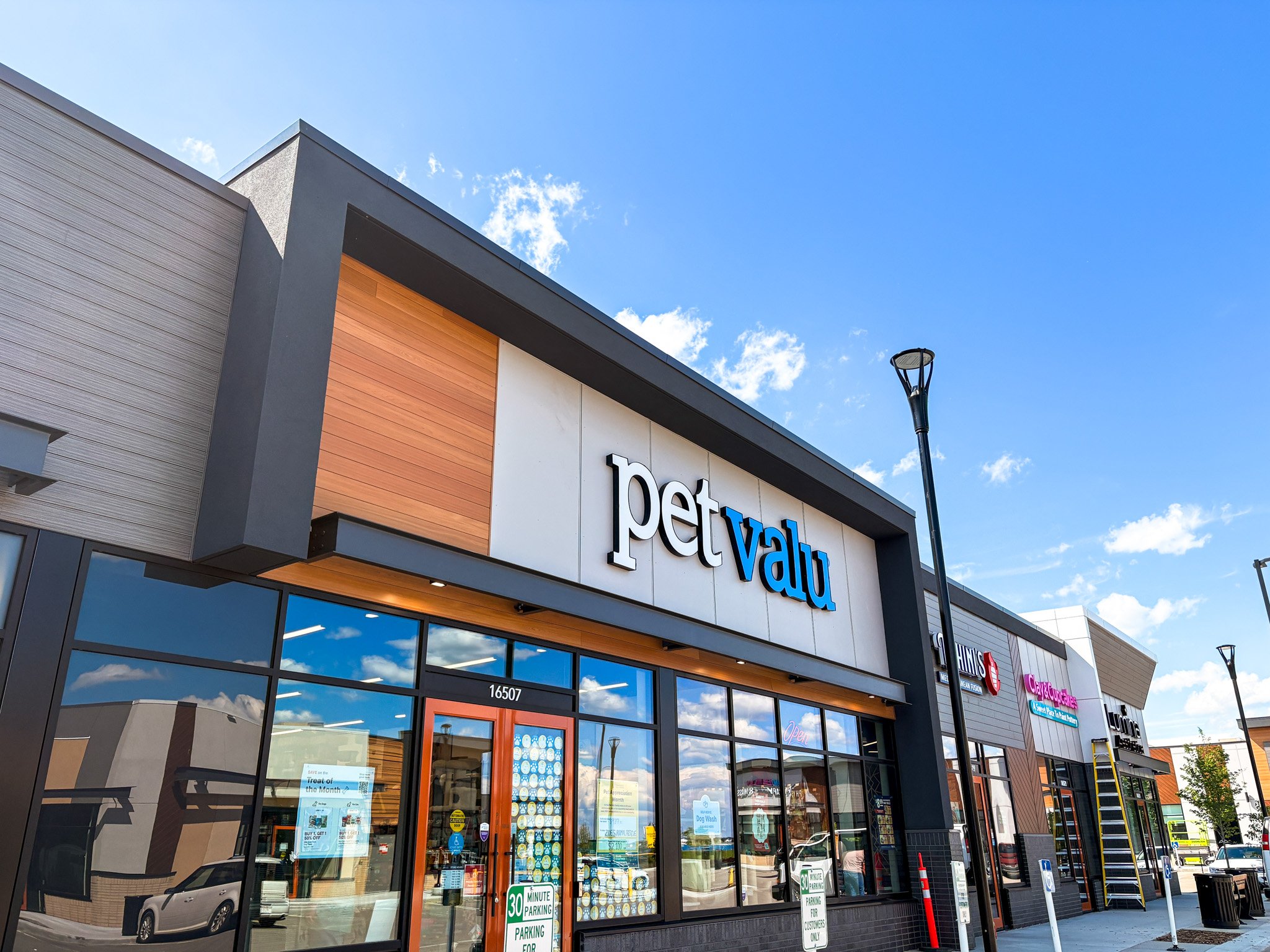 A modern retail store storefront with large glass windows and a sign that reads 'pet value'. The building has a combination of gray, orange, and white exterior panels. There is a parking lot in front with a few signs indicating parking restrictions. The sky is mostly clear with some clouds, and a streetlamp is visible.