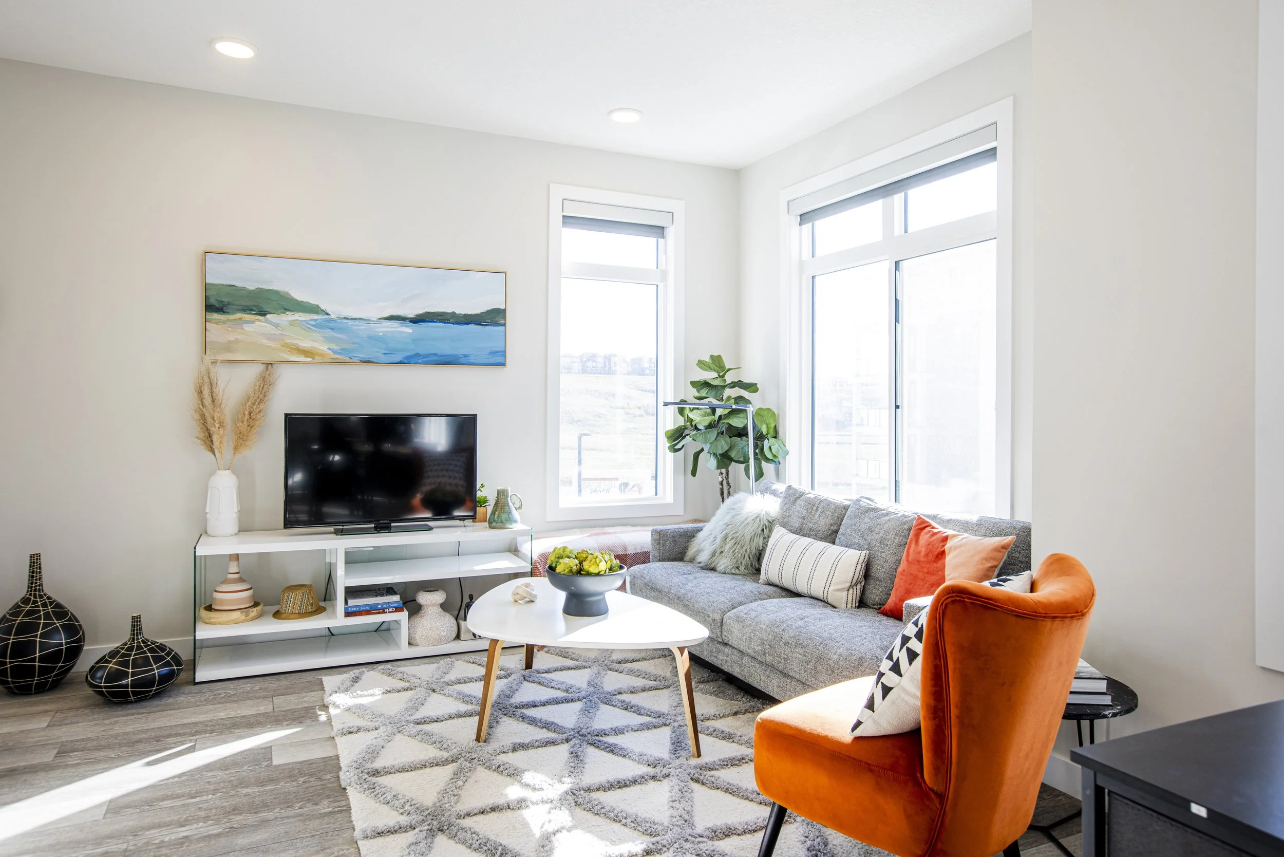 Bright living room with large windows, gray sofa with colorful pillows, orange armchair, white rug, modern white TV stand, flat screen TV, decorative vases, potted plant, and wall art displaying a landscape, natural light streaming in.
