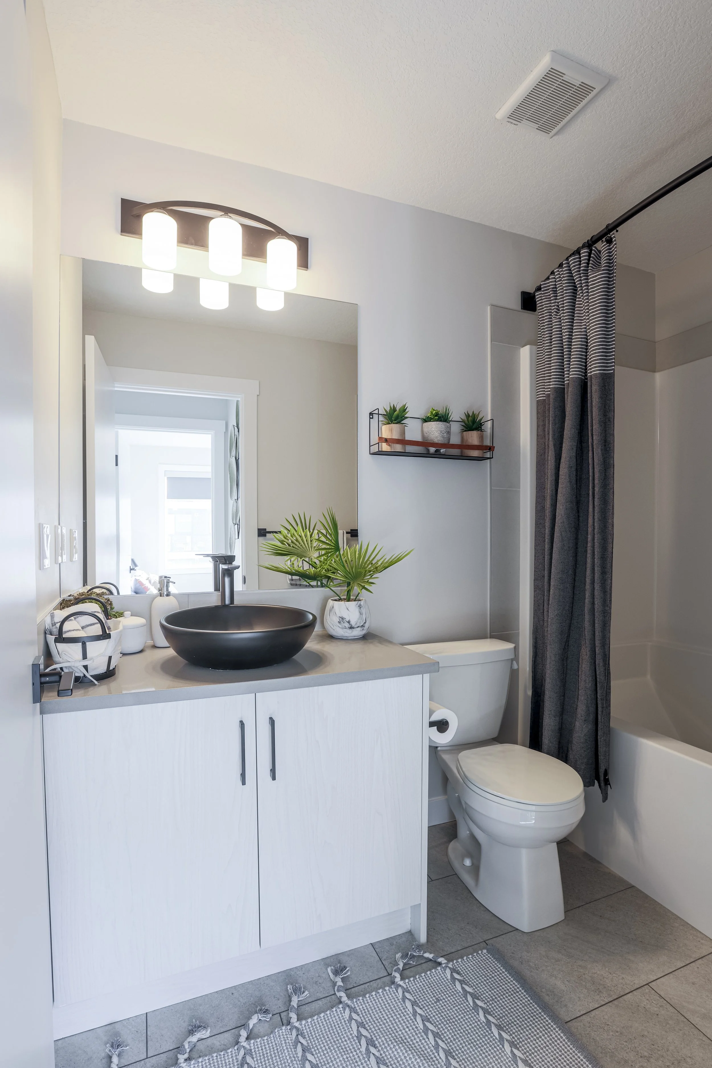 A modern bathroom with a white vanity, a black vessel sink, a large mirror, and a wall-mounted light fixture. There are three small potted plants on the shelf next to the mirror. A plant in a marble pot is on the vanity. The toilet is beside the vani