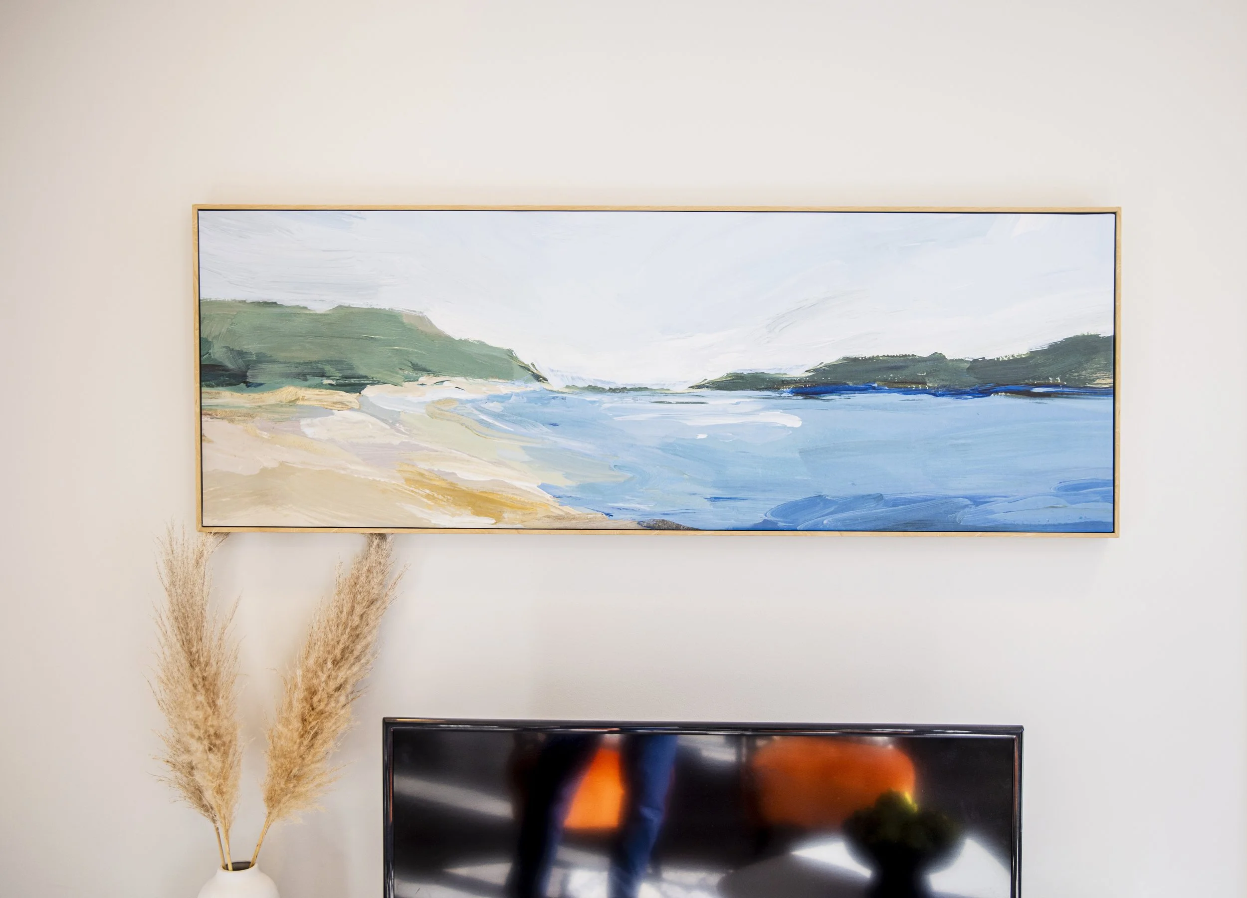 A large framed painting of a landscape with green hills, a beach, and a blue body of water on a beige wall. Below it, a television is partly visible, and to the left, vase with dried grasses.