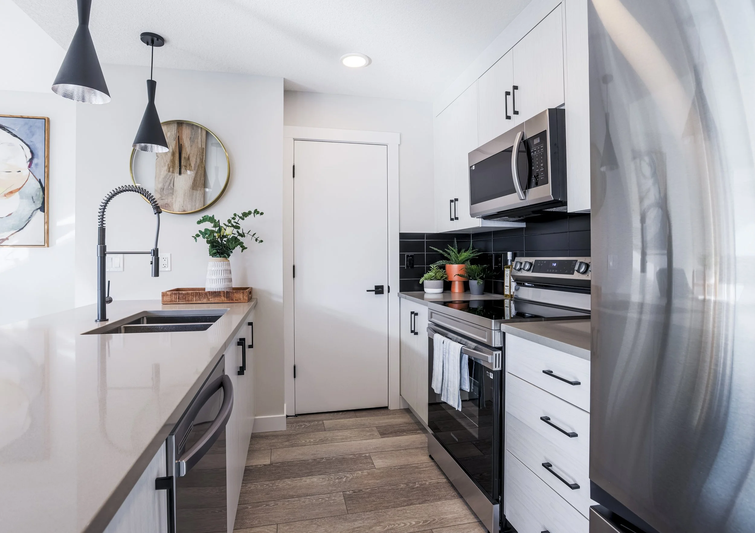 Modern kitchen with white cabinets, black backsplash, stainless steel appliances, a black oven, microwave, and fridge, with wooden flooring and decor including plants and artwork.