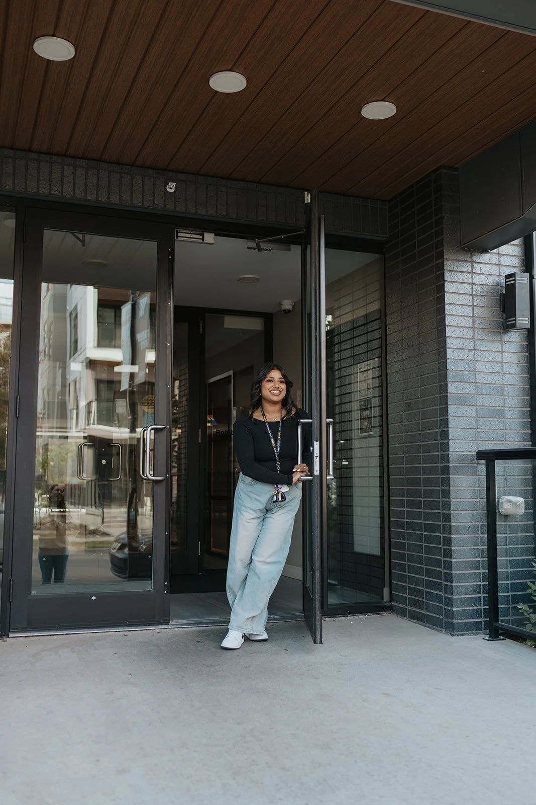 A woman standing in the doorway of a building, smiling and holding the door open, with a cityscape reflected in the glass behind her.