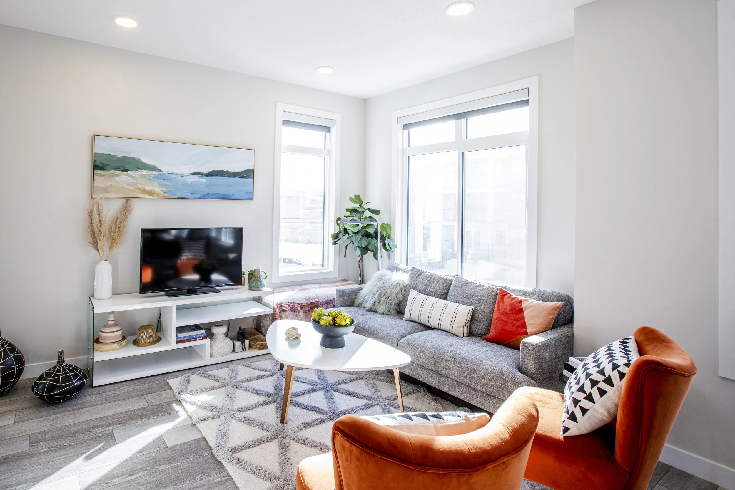Bright living room with a gray sofa, an orange armchair, a white coffee table, a TV on a white stand, large windows, a potted plant, and decorative vases.