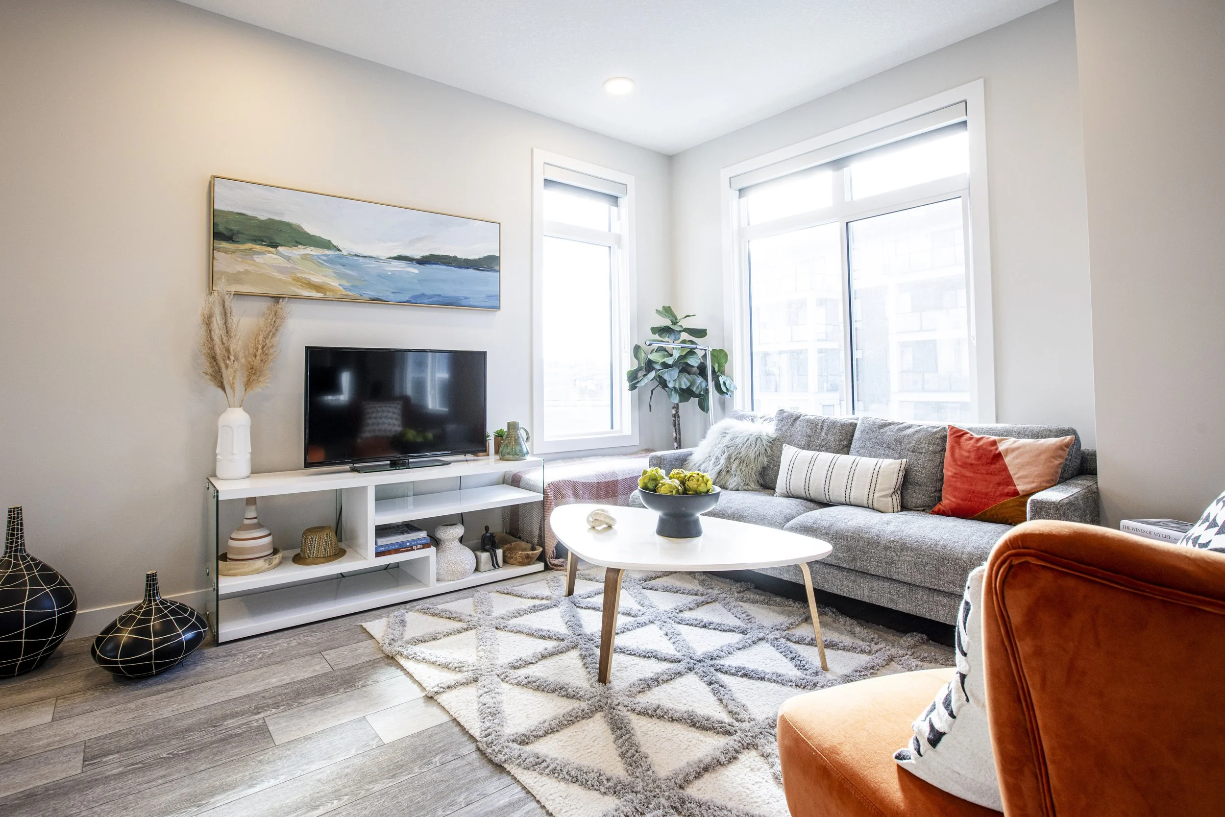 Modern living room with grey sofa, orange armchair, white coffee table, and large windows letting in natural light.