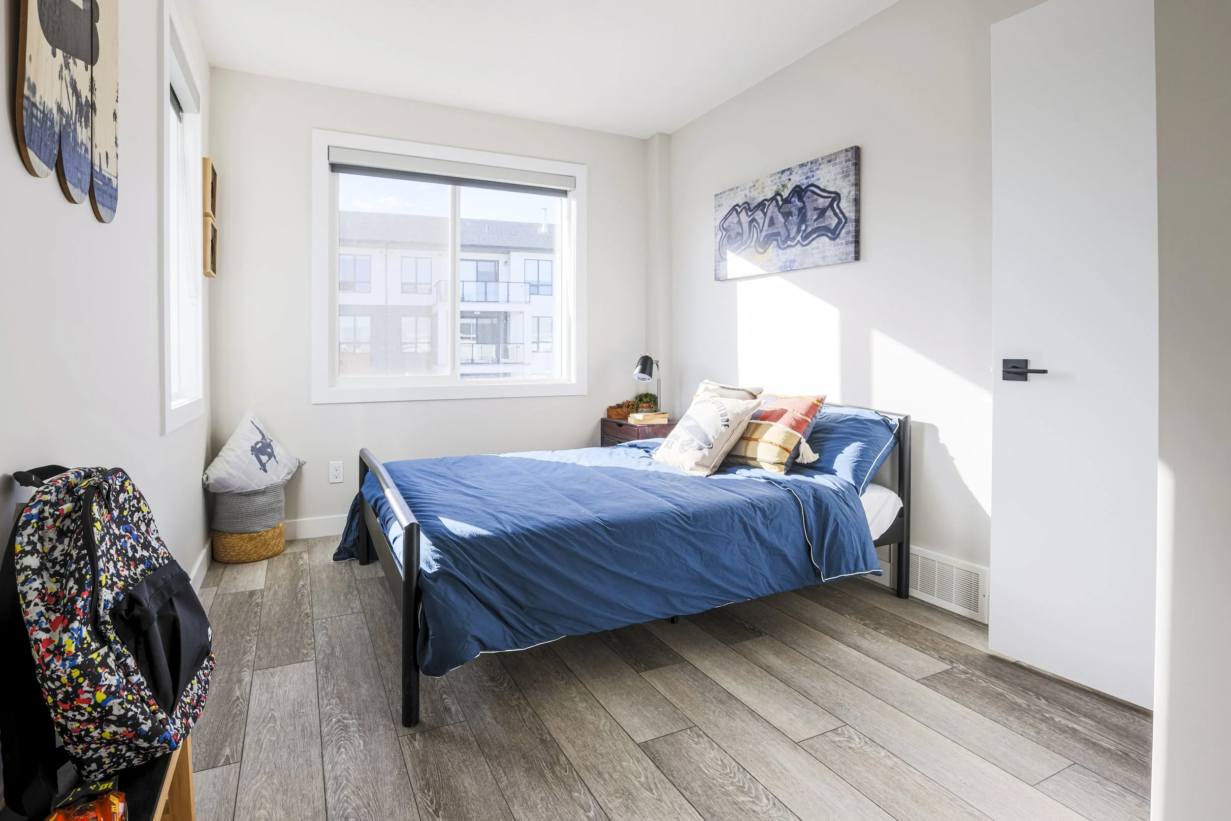 A clean, bright bedroom with a large window, a bed with blue bedding, a small nightstand with a lamp, a backpack on the floor, and modern decor on the walls.