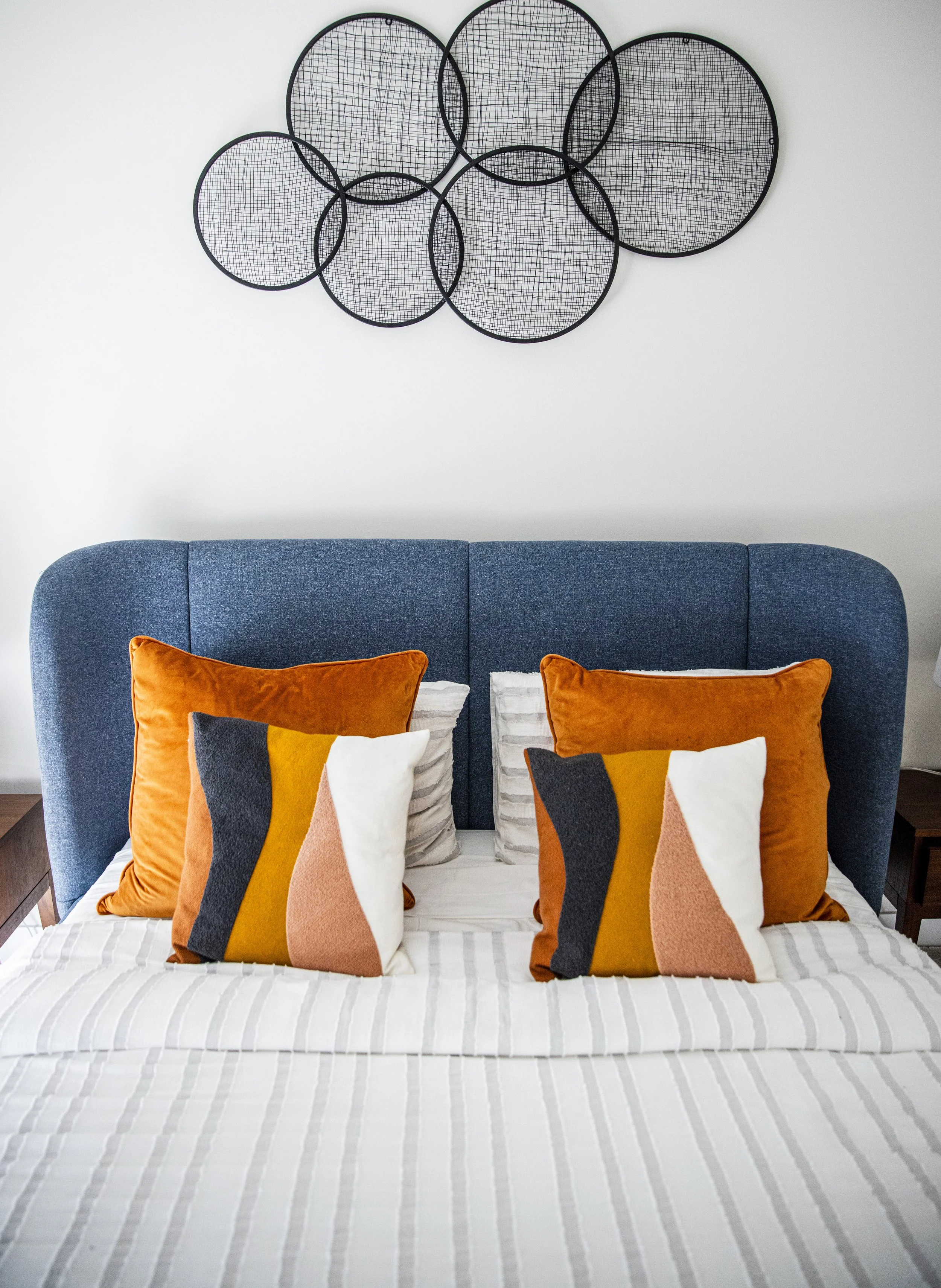 A modern bedroom with a blue upholstered headboard, decorated with four colorful pillows featuring abstract patterns in orange, mustard yellow, pink, white, and black. Above the bed, there is a wall art piece comprising several black wire circles of 