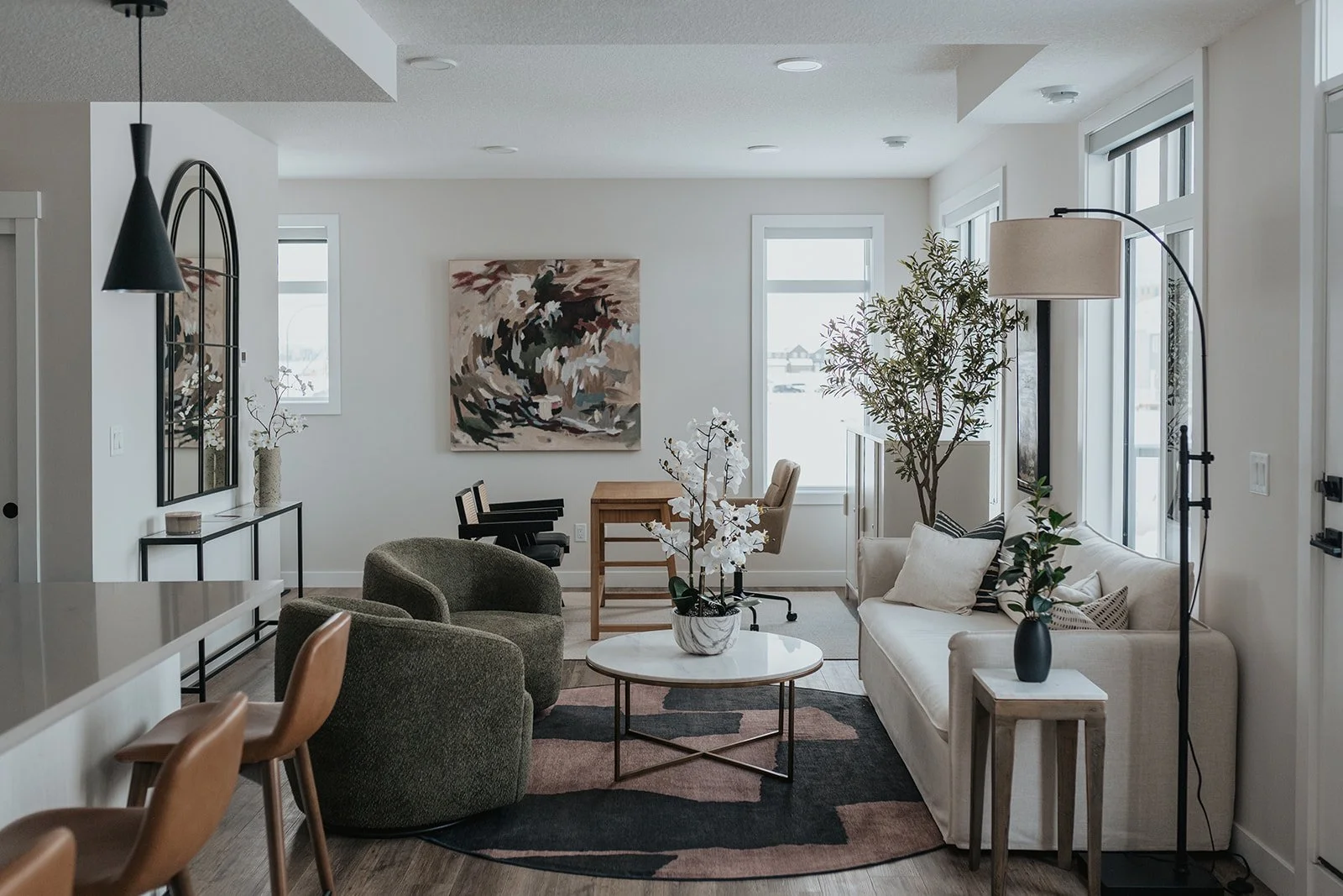 Living room with white walls, large windows letting in natural light, a white couch with pillows, a coffee table with a vase of white flowers, dark green armchairs, a black floor lamp, a side table, a large abstract painting on the wall, and plants.