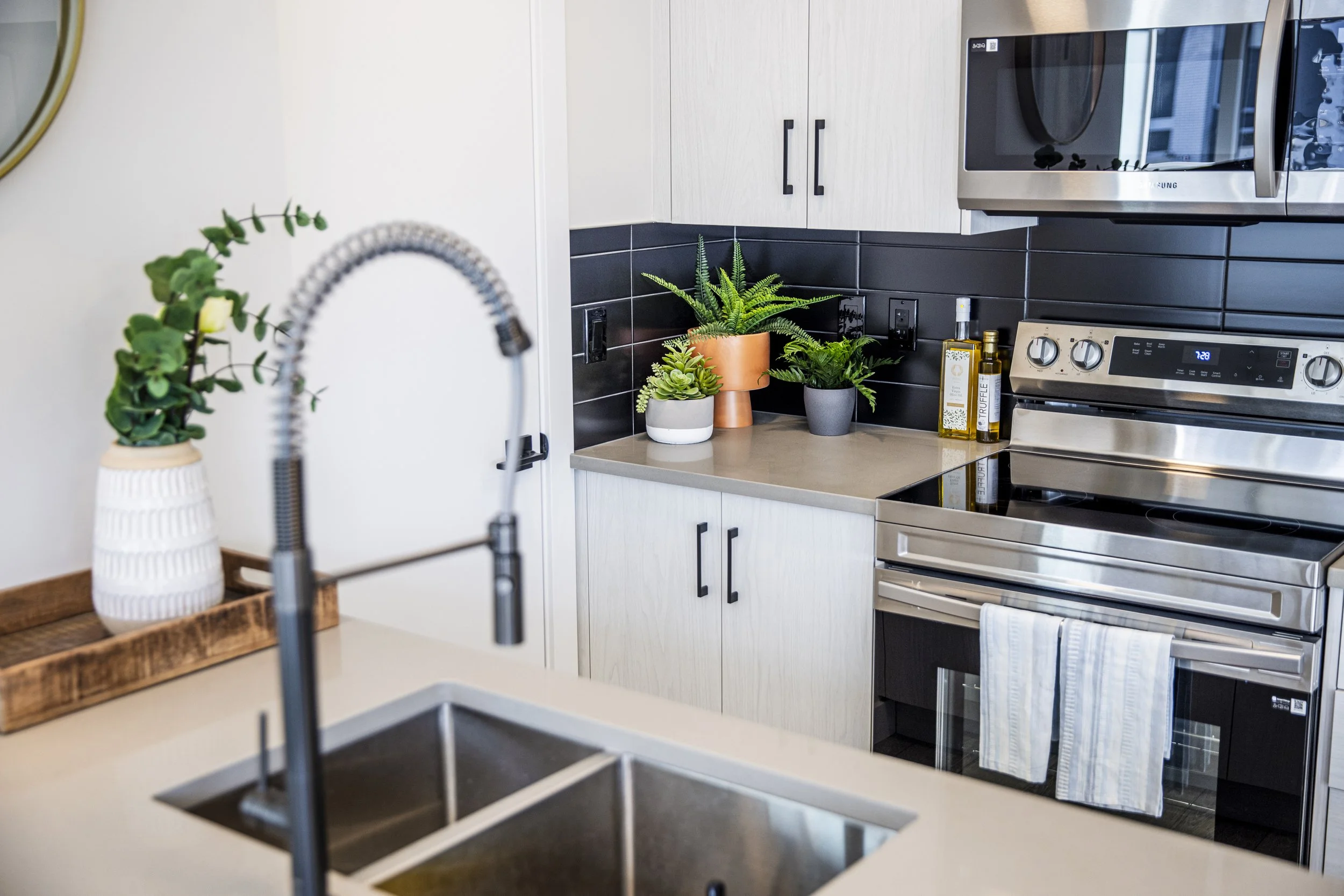 Kitchen with white cabinets, black backsplash, stainless steel stove, microwave, and a beige countertop decorated with potted green plants.