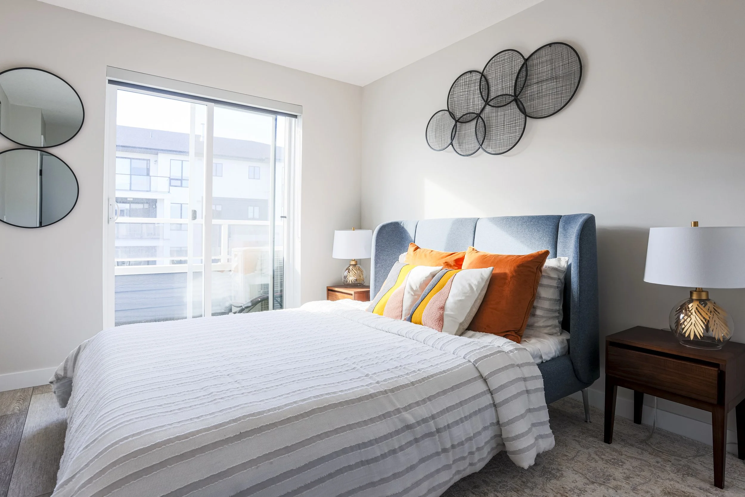 Modern bedroom with a large bed, colorful pillows, bedside lamps, sliding glass door, and decorative wall art.