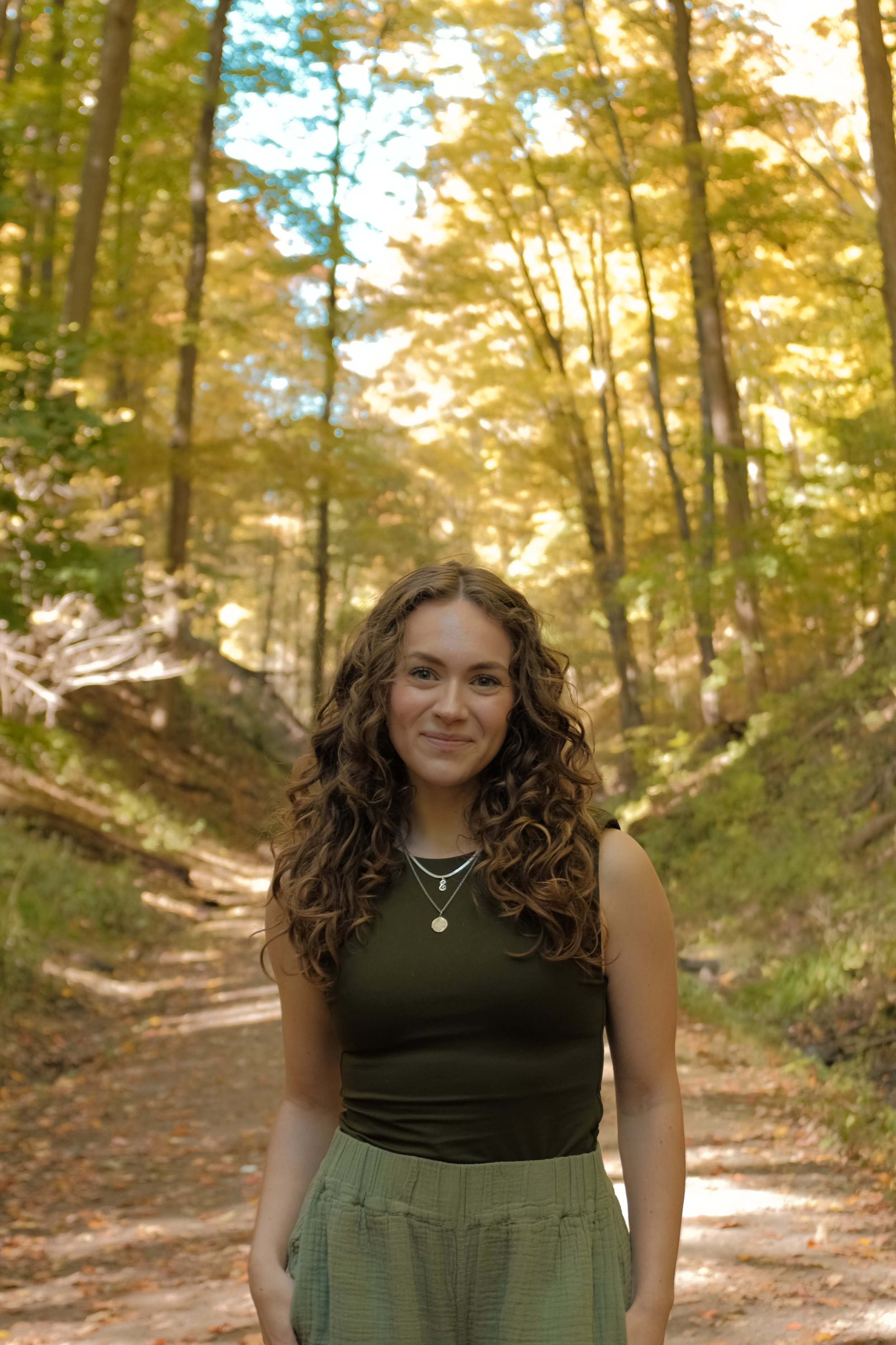Emily Grunthal, Founder of Noia Wellness, smiling in a green tanktop and green pants standing in front of a forest with tall trees and yellow leaves