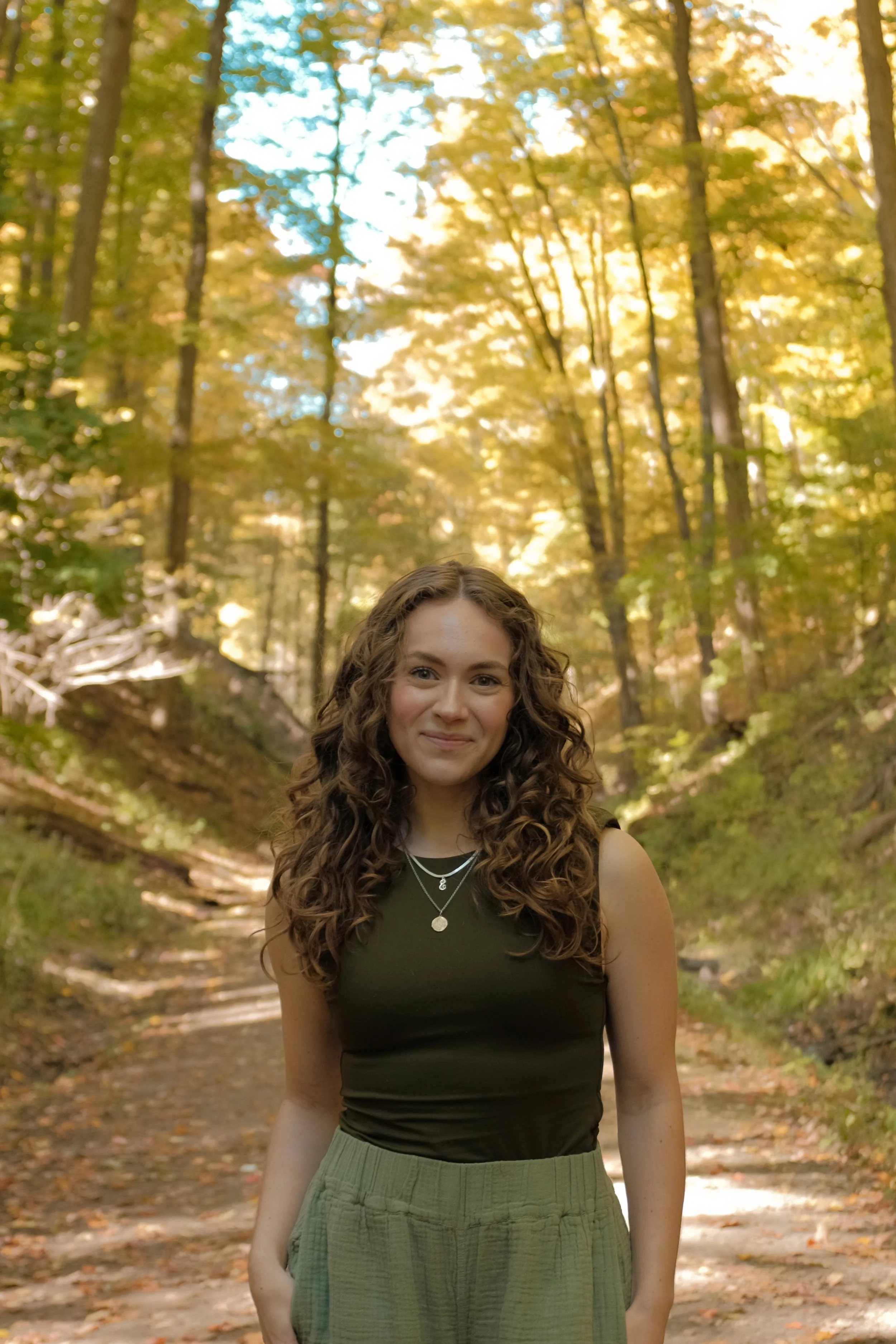 Founder of Noia Wellness, Emily GRunthal, smiling standing on a forest path in Autumn with yellowing leaves behind her. She is wearing a dark green top with curly hair and visible gold necklaces.