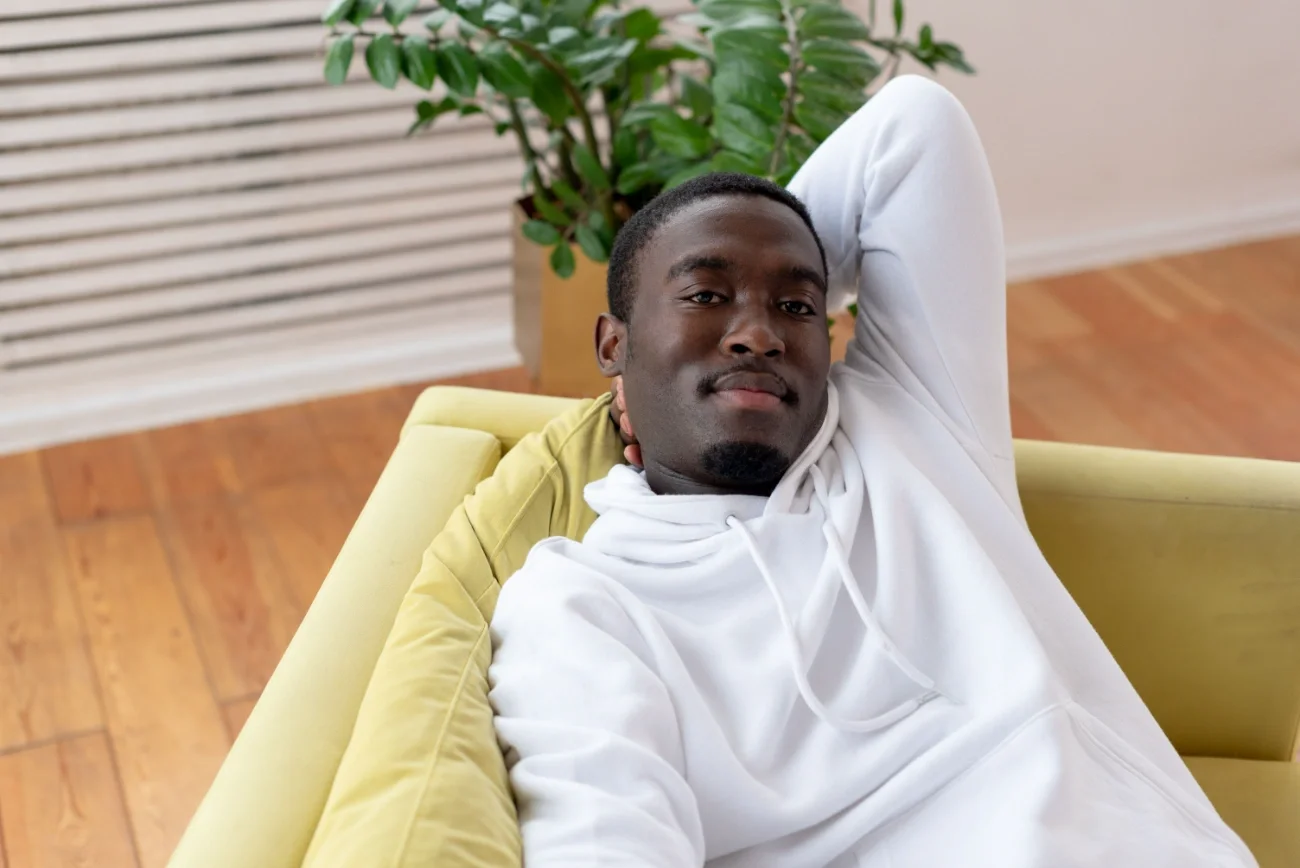 A man sitting on a light green couch with his hand behind his head, smiling at the camera.