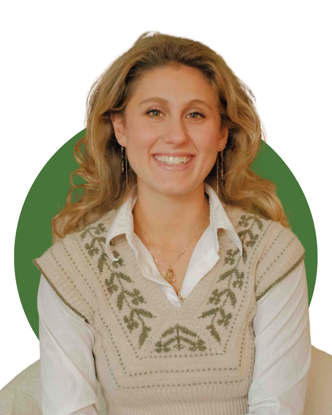 A woman with wavy blonde hair smiling and sitting on a beige armchair in a cozy living room, wearing a white shirt and a patterned sweater vest.