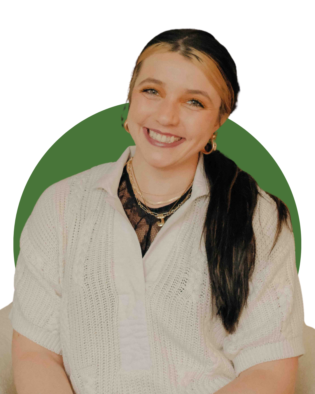 A woman with long dark hair with blond streaks, smiling while sitting on a cream-colored armchair in a cozy living room. Behind her is a wooden mantle, decorated with framed pictures, plants, and small decorative items. The wall above has three framed landscape pictures, and the room has warm lighting.