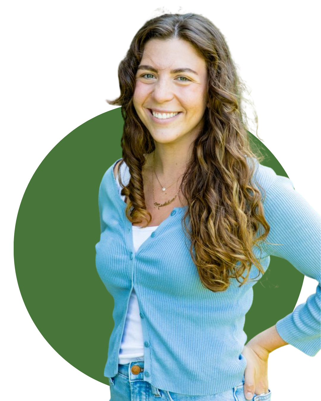  with long, curly brown hair smiling and sitting on a white armchair in a cozy living room. Behind her is a wooden fireplace mantel with decor items, including a framed photo, a small potted plant, and a decorative owl. There are three framed portraits of landscapes or abstract art hanging on the wall above. To the right, there is a large green leafy plant and a small side table with more plants and objects.