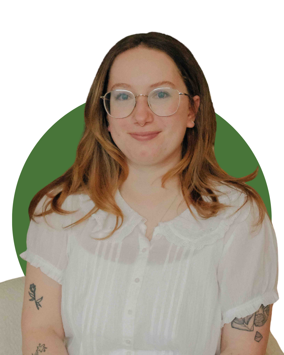 A woman sitting on a cream-colored chair in a living room, smiling at the camera. She has shoulder-length light brown hair, wears glasses, a white button-up shirt, and olive green pants. She has tattoos on her arms and is seated in front of a dark wooden fireplace mantel with decorative items and framed pictures, and there is a large green plant behind her.