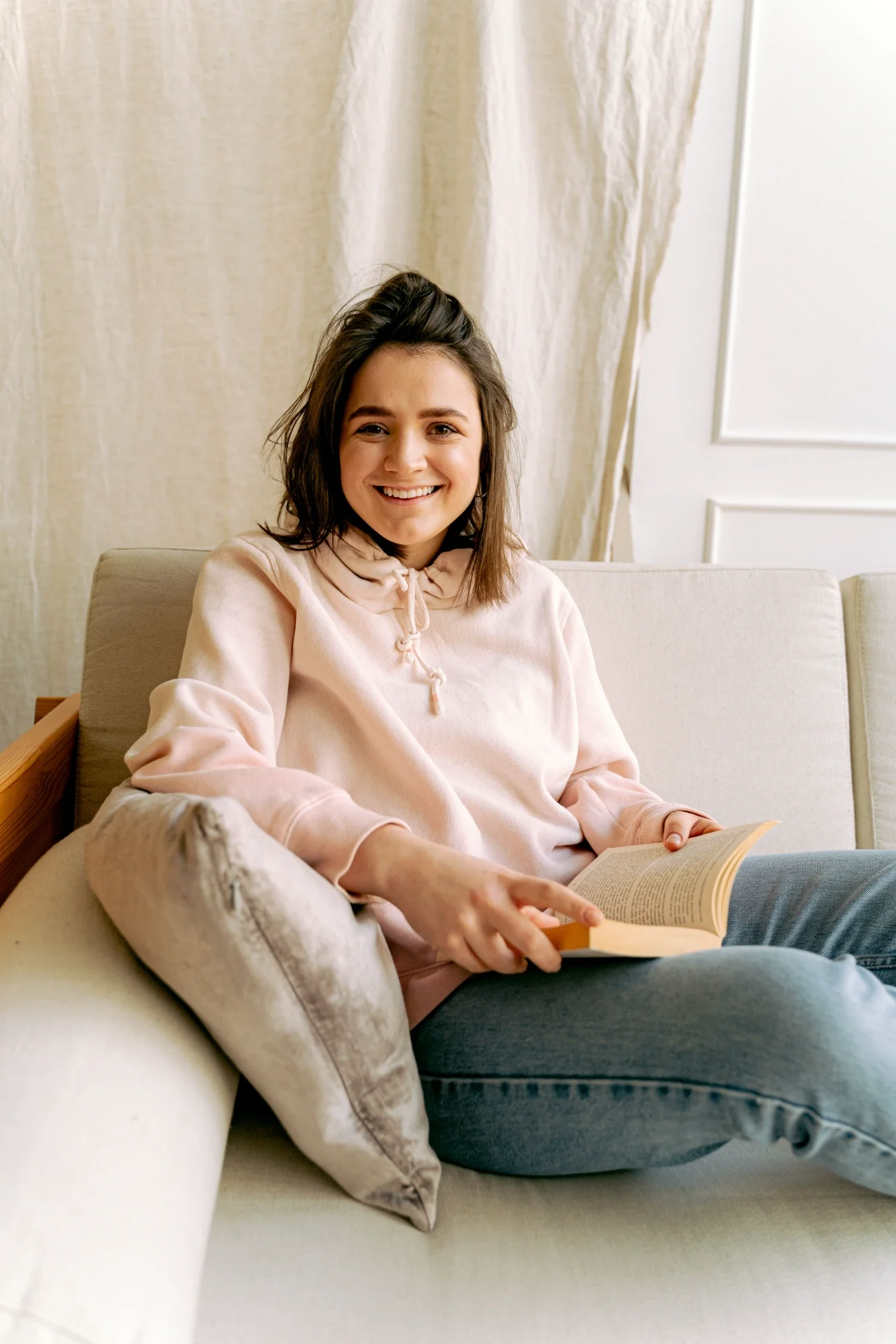 A young woman sitting on a couch reading a book, smiling at the camera.