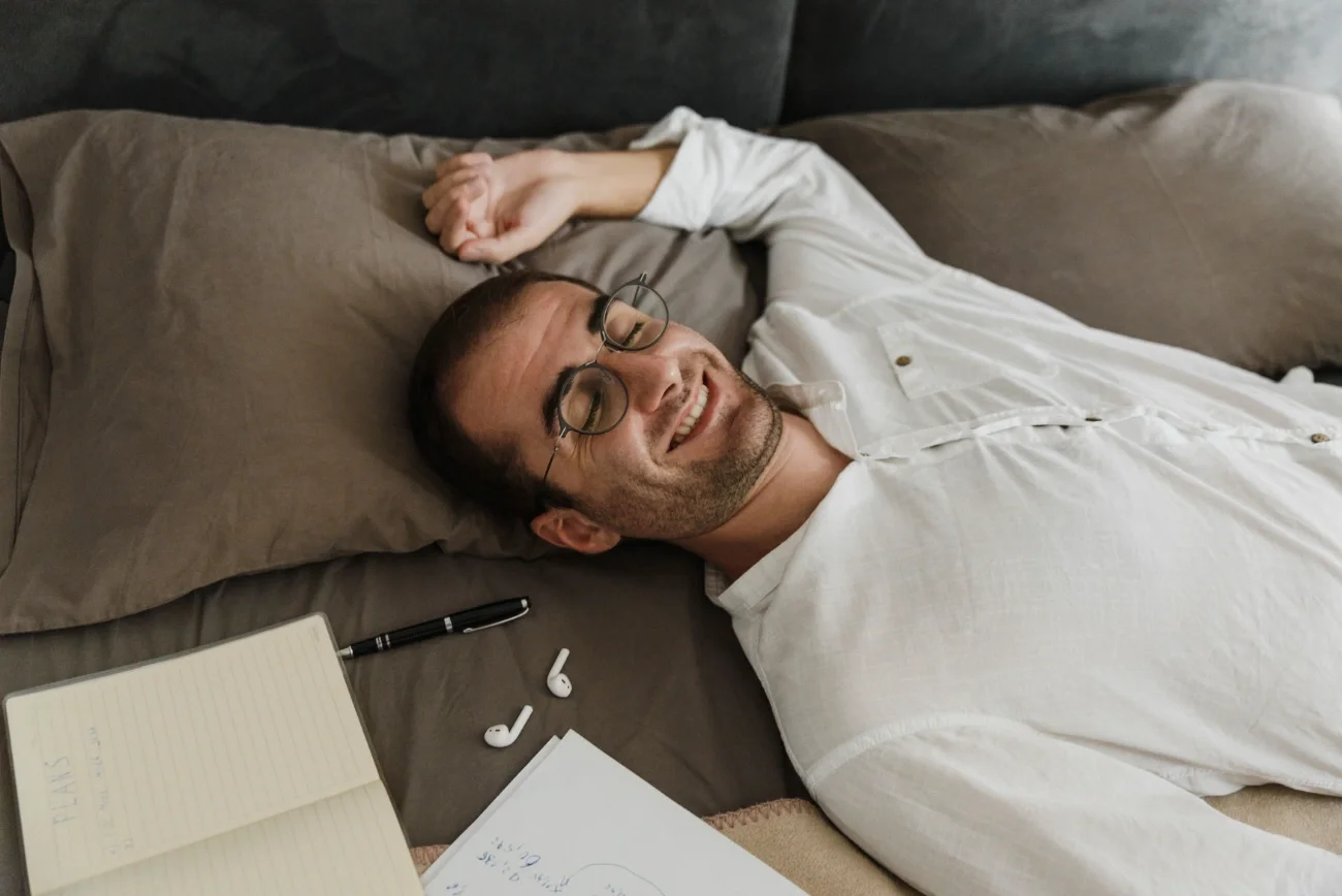 A man laying down on a bed surrounded by airpods and a notebook.