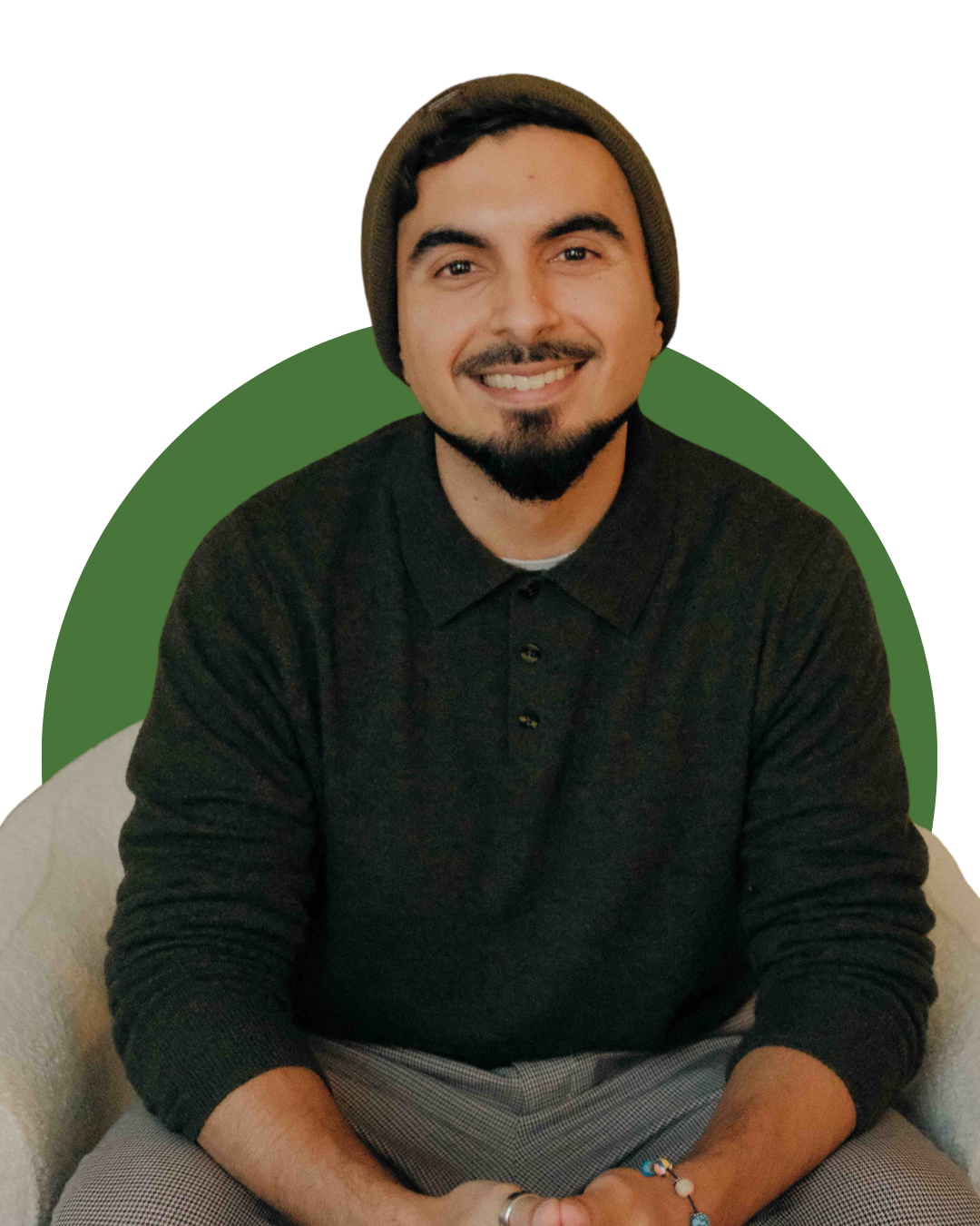 A man with dark hair, a beard, and a mustache, wearing a gray beanie, black sweater, gray plaid pants, sitting on a white armchair, smiling at the camera in a cozy living room with a wooden fireplace mantel, green houseplants, framed pictures, and decorative items.