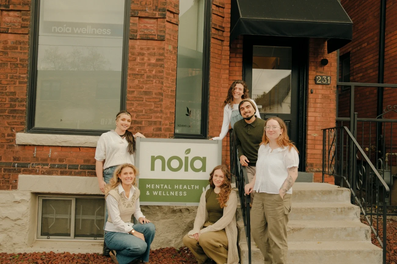 The Noia Wellness team standing in front of their Locke Street South office, standing and sitting around a sign featuring their logo.